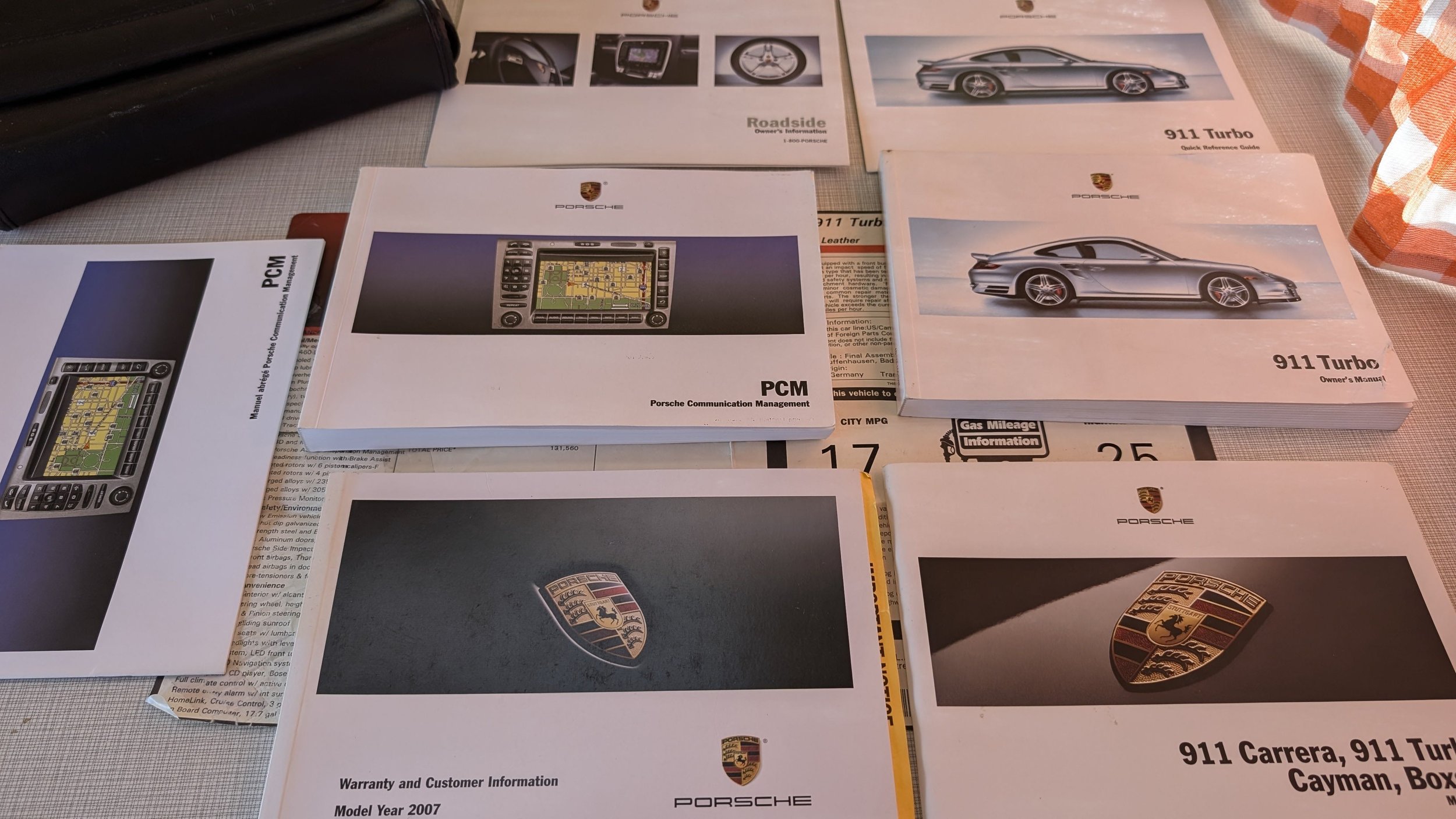 Porsche car manuals and information booklets, including the owner’s manual and communication management guides, spread out on a table with some documents and a curtain on the side.