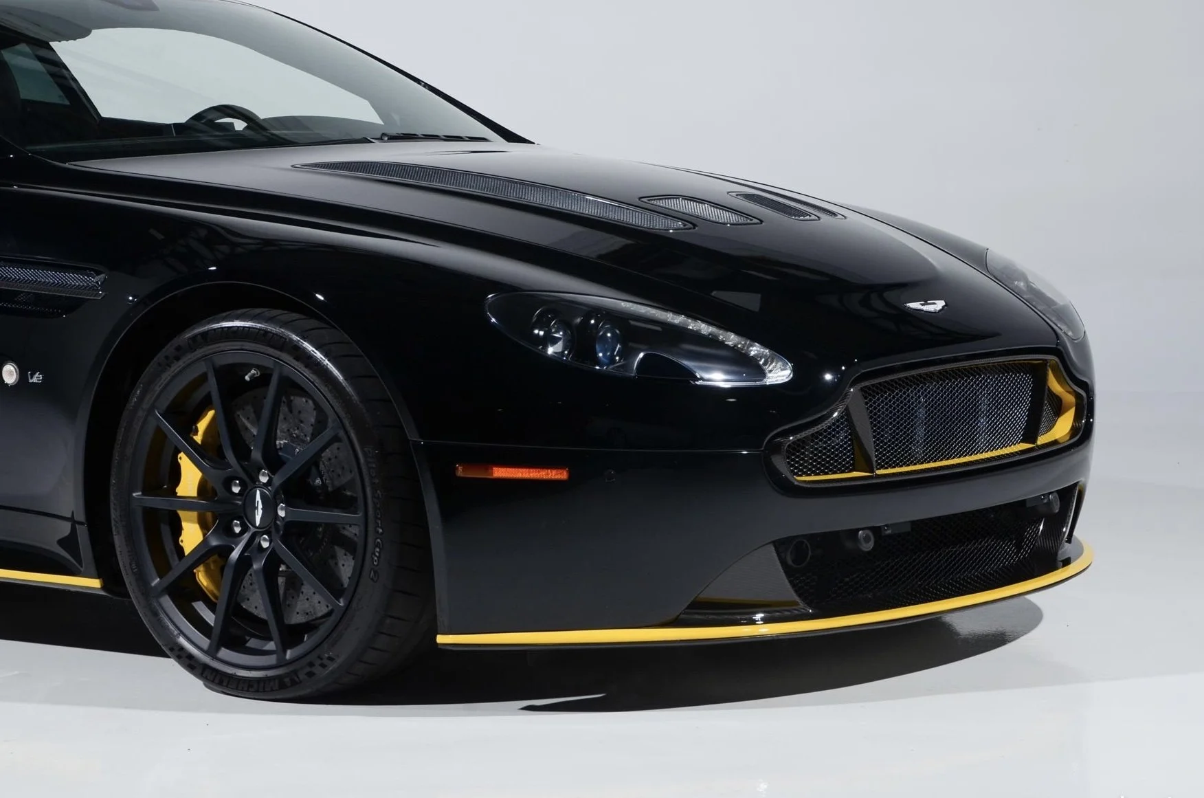 Close-up of a black sports car with yellow accents, showing front left wheel, headlight, and grille with mesh design.