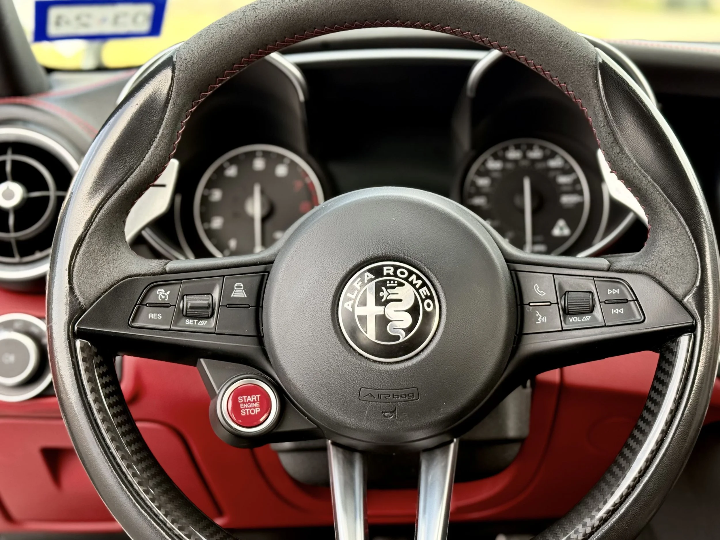 Close-up of a steering wheel inside an Alfa Romeo car, featuring buttons for cruise control, volume, calling, and voice commands, with a red start/stop engine button below.