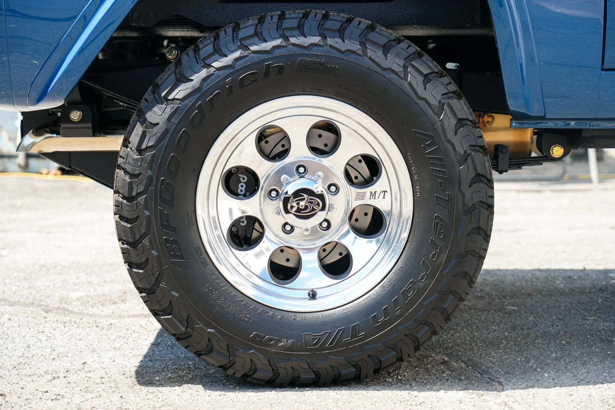 Close-up of a rugged off-road tire on a vehicle, with a shiny silver rim and multiple circular cutouts, mounted on a blue vehicle.