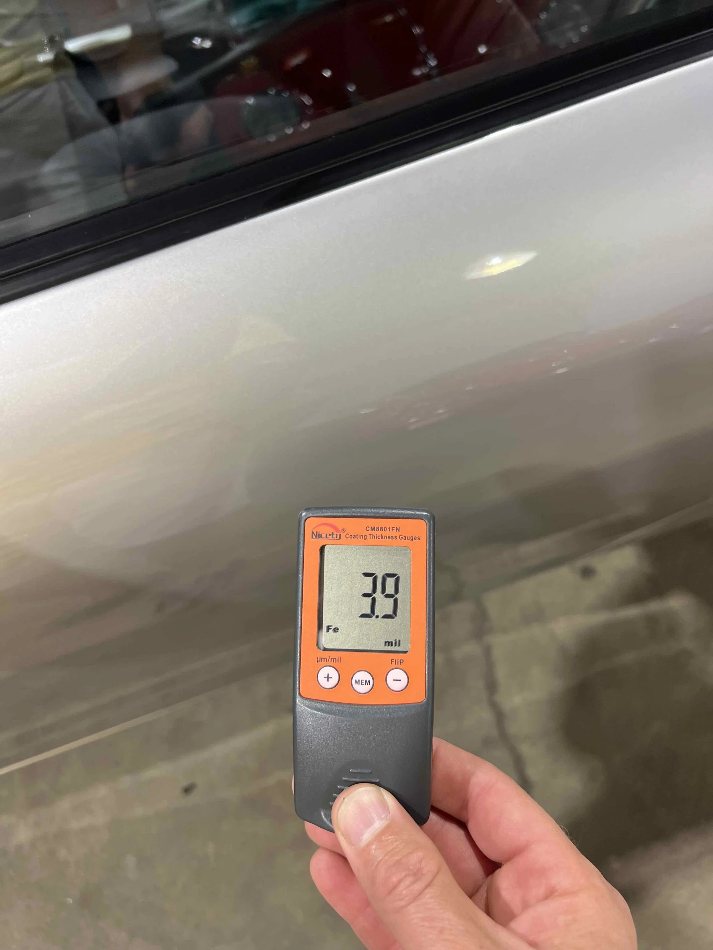 Close-up of a hand holding a coating thickness gauge measuring a silver-colored car door, displaying a reading of 3.9 mil.
