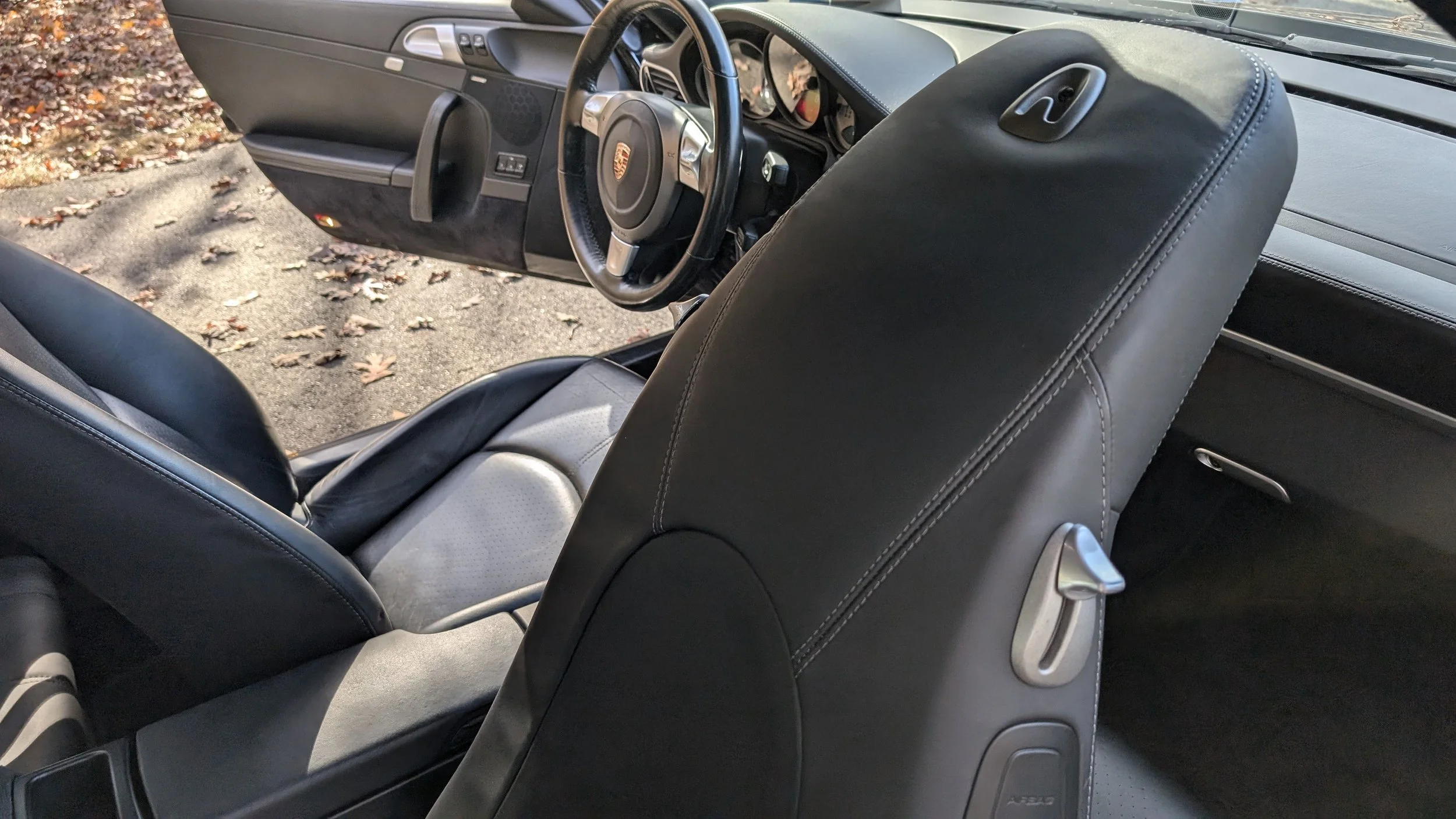 Interior view of a Porsche vehicle showing the black leather front seats, steering wheel, and dashboard with visible control buttons and gauges, parked outside with fallen leaves on the ground.
