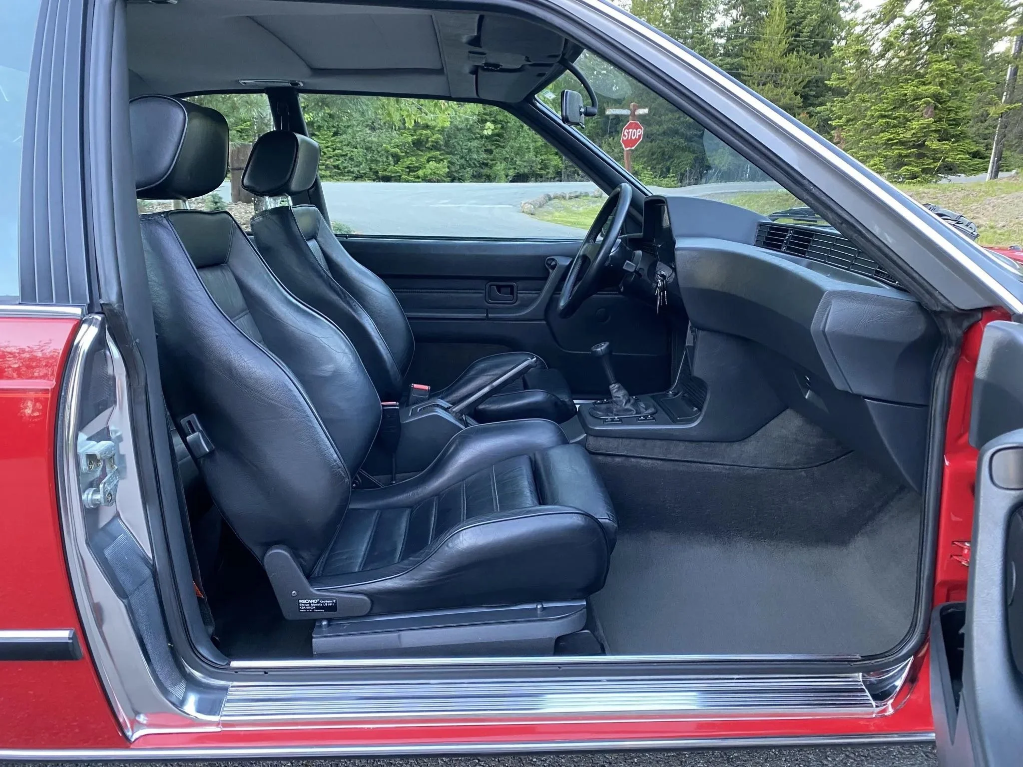 The front interior of a red vintage car featuring black leather seats, an automatic gear shift, and a black dashboard, with a view of a road and a stop sign outside.