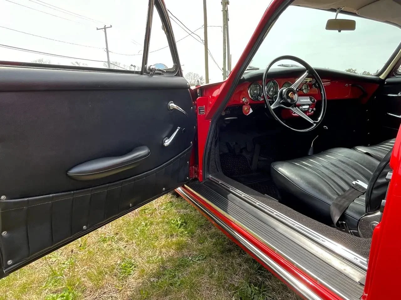 Interior of a vintage red car showing the black door panel and dashboard, with a black seat, steering wheel, and speedometer gauges.