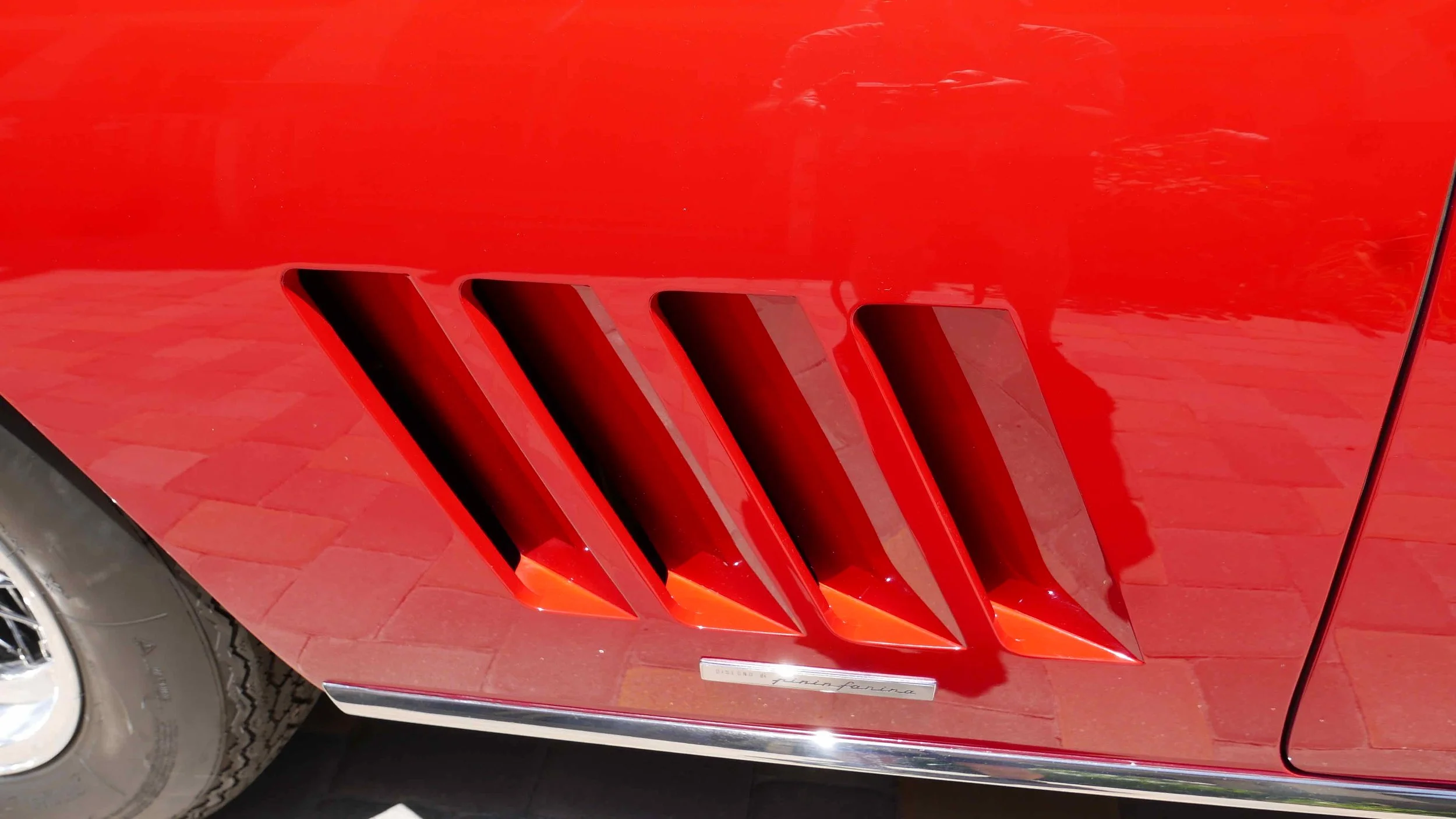 Close-up of a red classic car's side vent with four angled, black cooling openings.