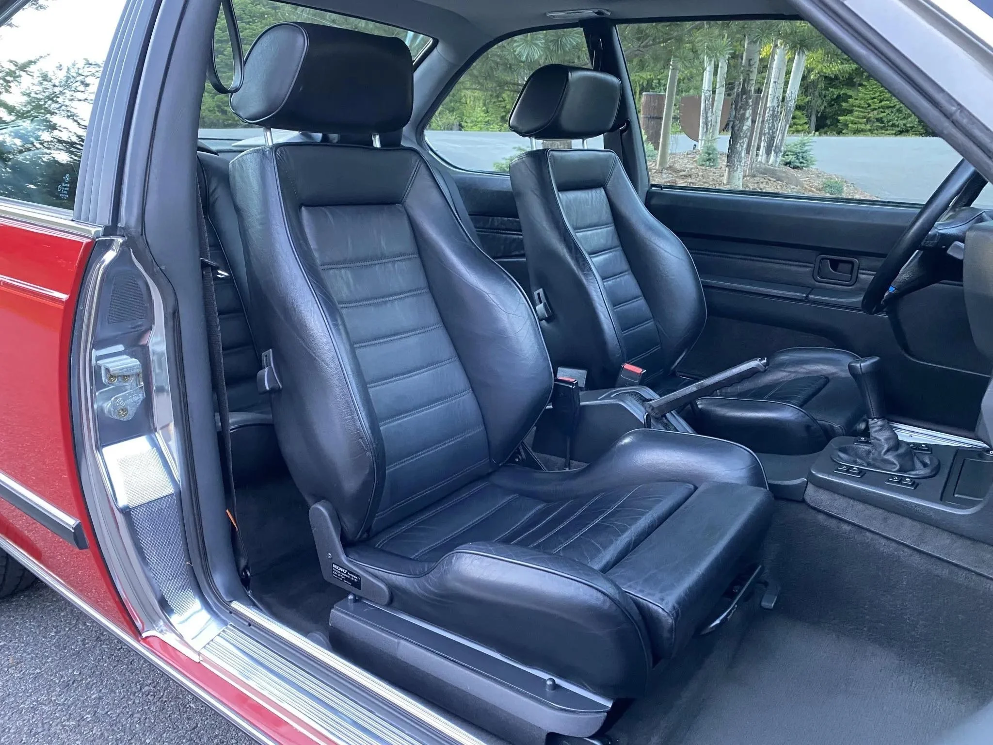 The interior of a vintage car showing two black leather seats, a gear shift, and a dashboard with a window view of trees outside.
