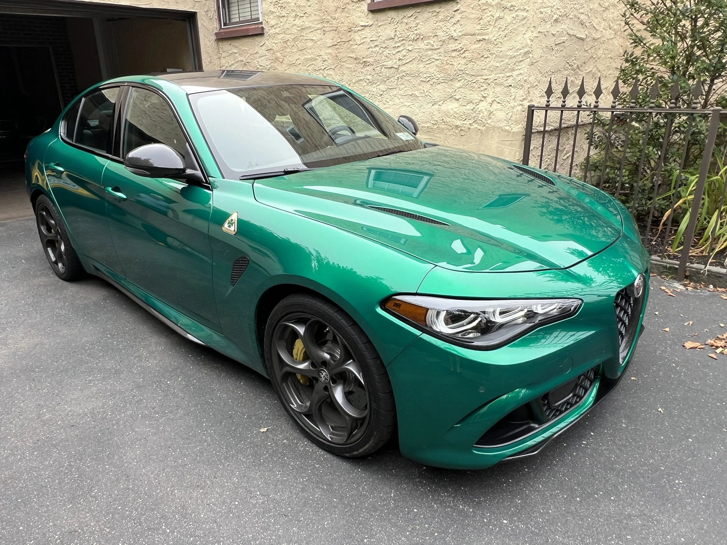Green Alfa Romeo sports car parked on driveway next to a beige textured wall, with dark alloy wheels and silver side mirror.