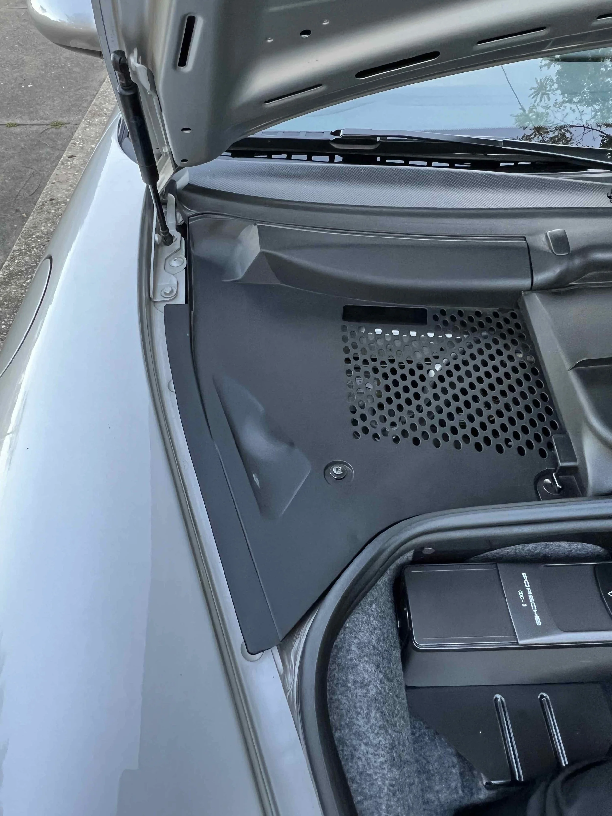 Open trunk of a silver Porsche sports car showing a black plastic vented cover and a black Porsche branded electronic device inside, with part of the car's exterior and windshield visible.