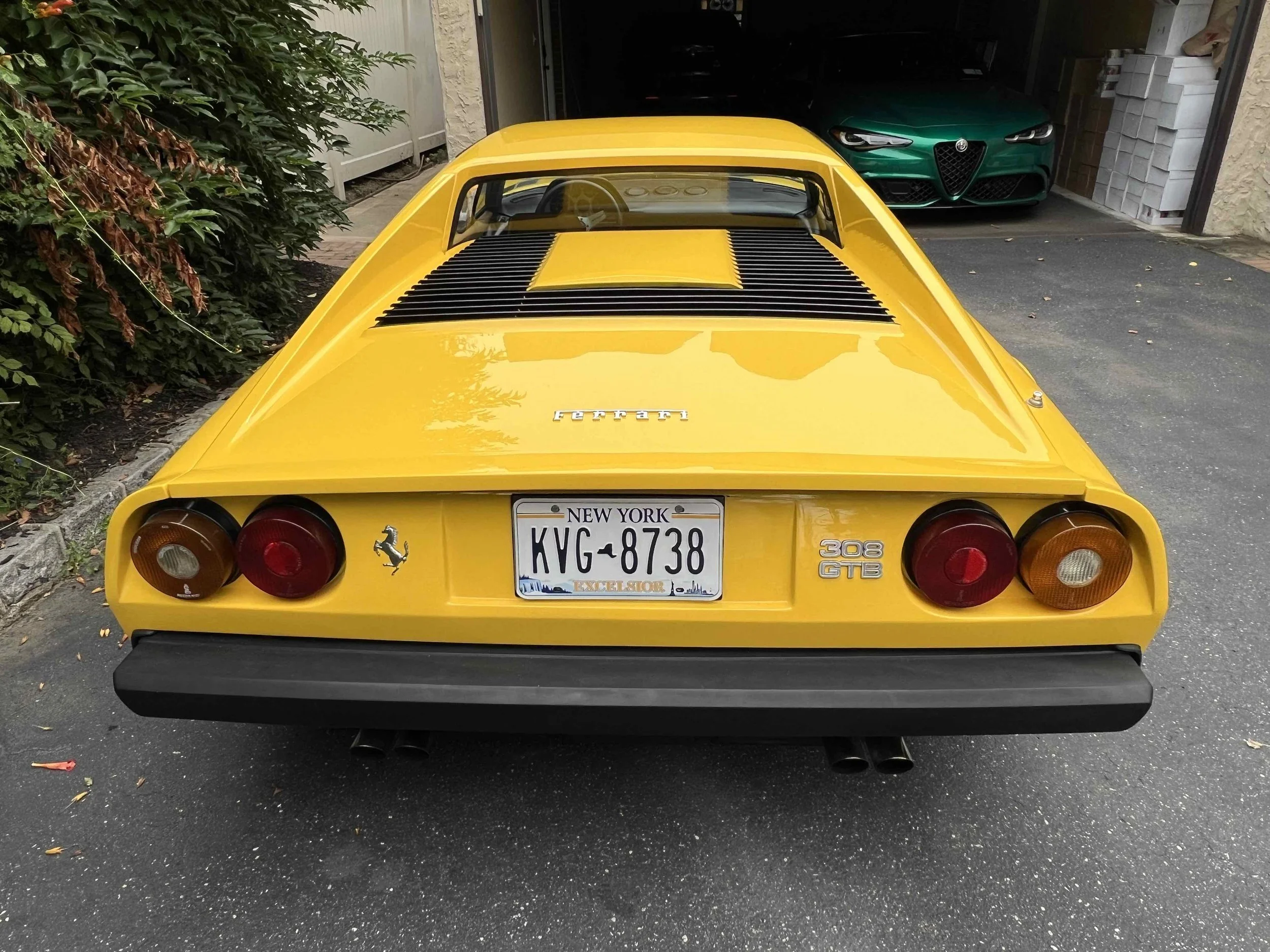 Yellow Ferrari 308 GTB parked in driveway, with a New York license plate, and a green Alfa Romeo parked inside garage in the background.