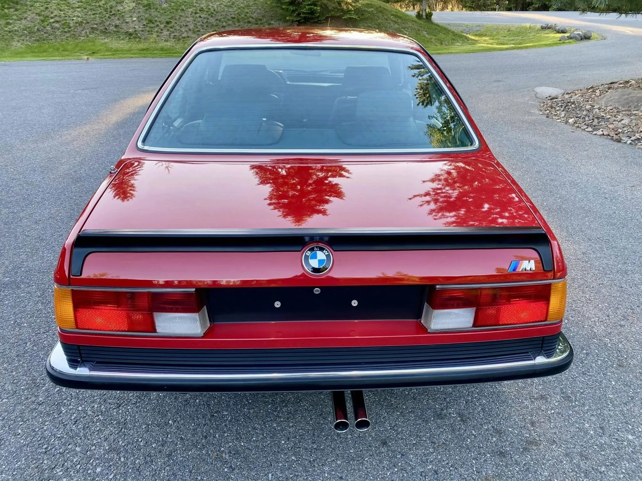 Red vintage BMW M car with dual exhaust pipes, parked on a gravel surface, reflecting trees and sky in the rear window.