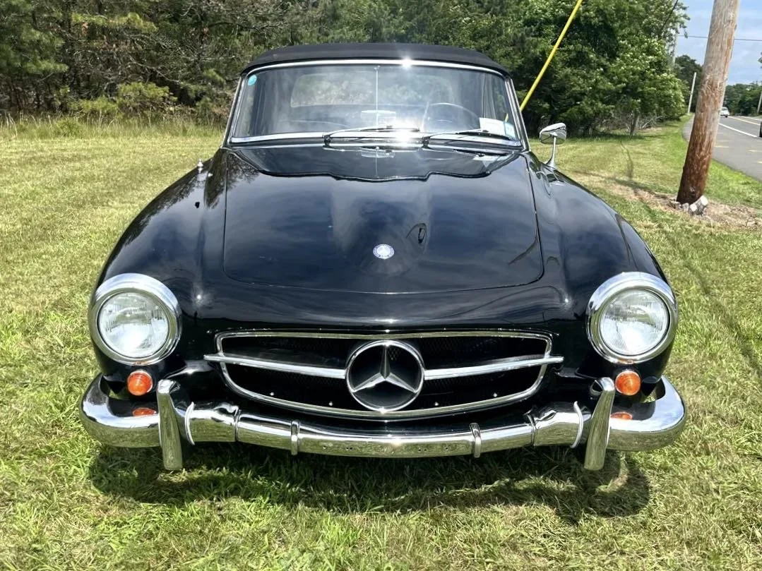 A black vintage Mercedes-Benz car parked on grass near roadside with trees in the background.