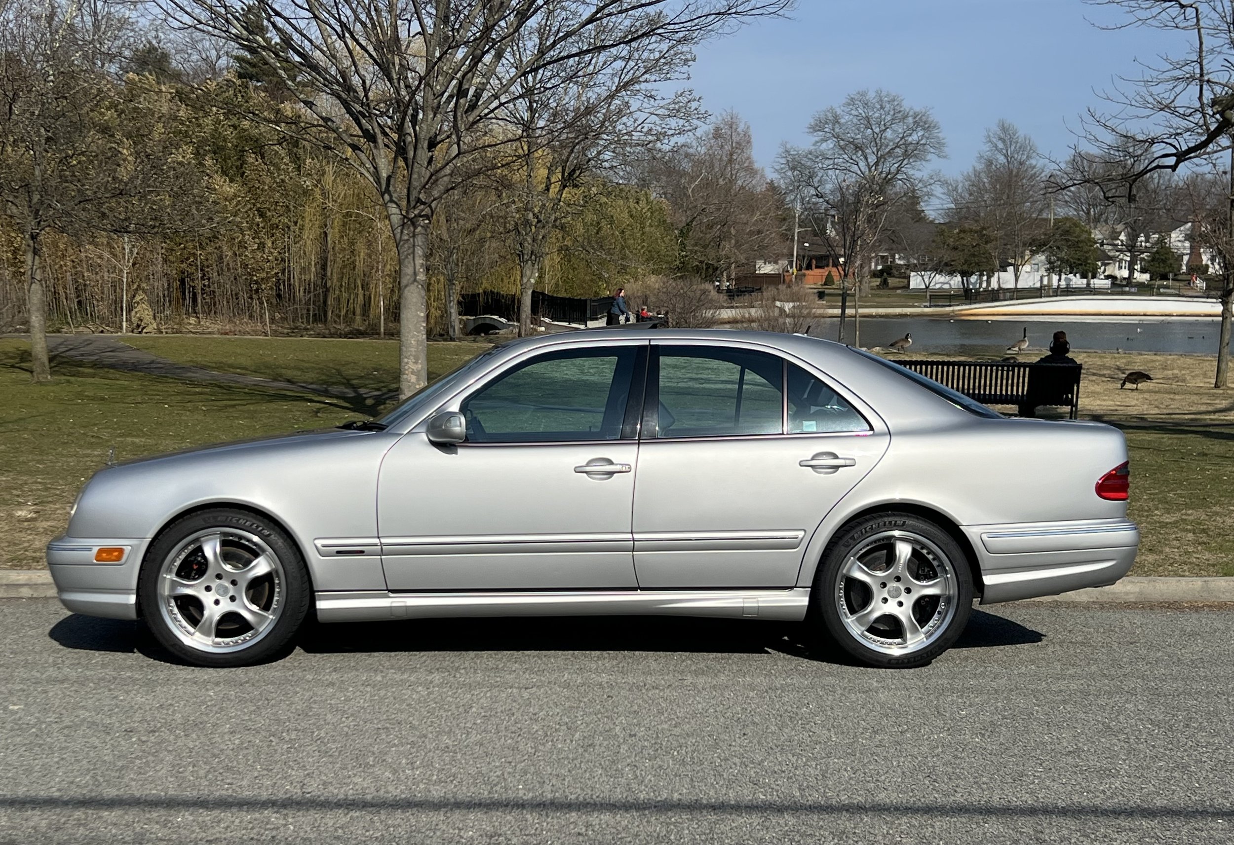 A silver Mercedes-Benz sedan parked on the street with a park, trees, a pond, and houses in the background.