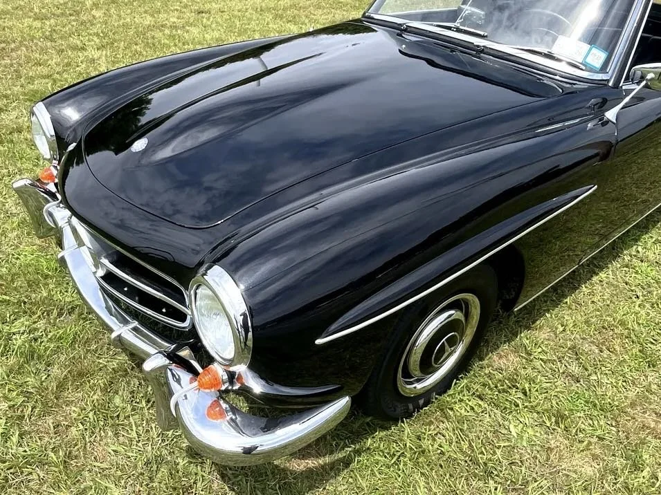 Close-up of a classic black car, likely a vintage model, parked on grass, showing its front left side including headlight, bumper, and side detail.