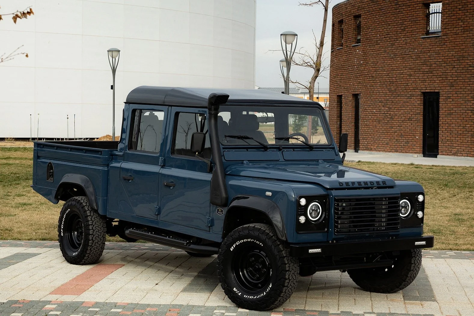 A blue Land Rover Defender pickup truck parked on a paved surface with grassy area and modern buildings in the background.