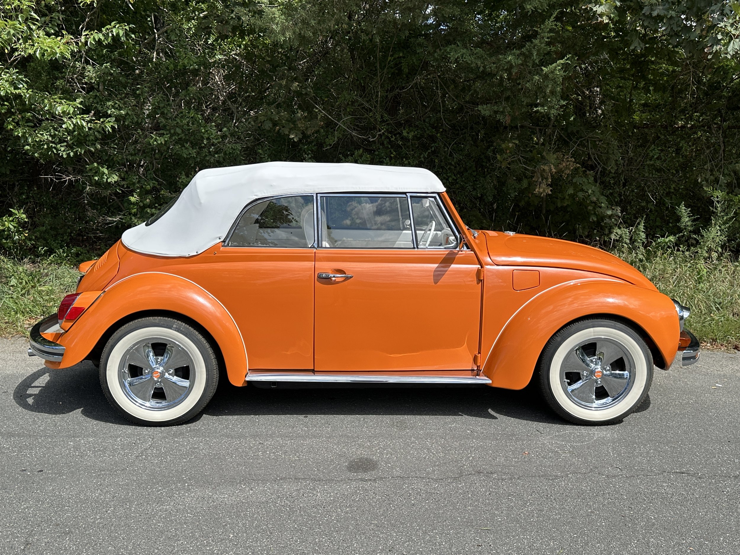Side view of a vintage orange Volkswagen Beetle convertible with a white soft top, parked on a paved road with greenery in the background.