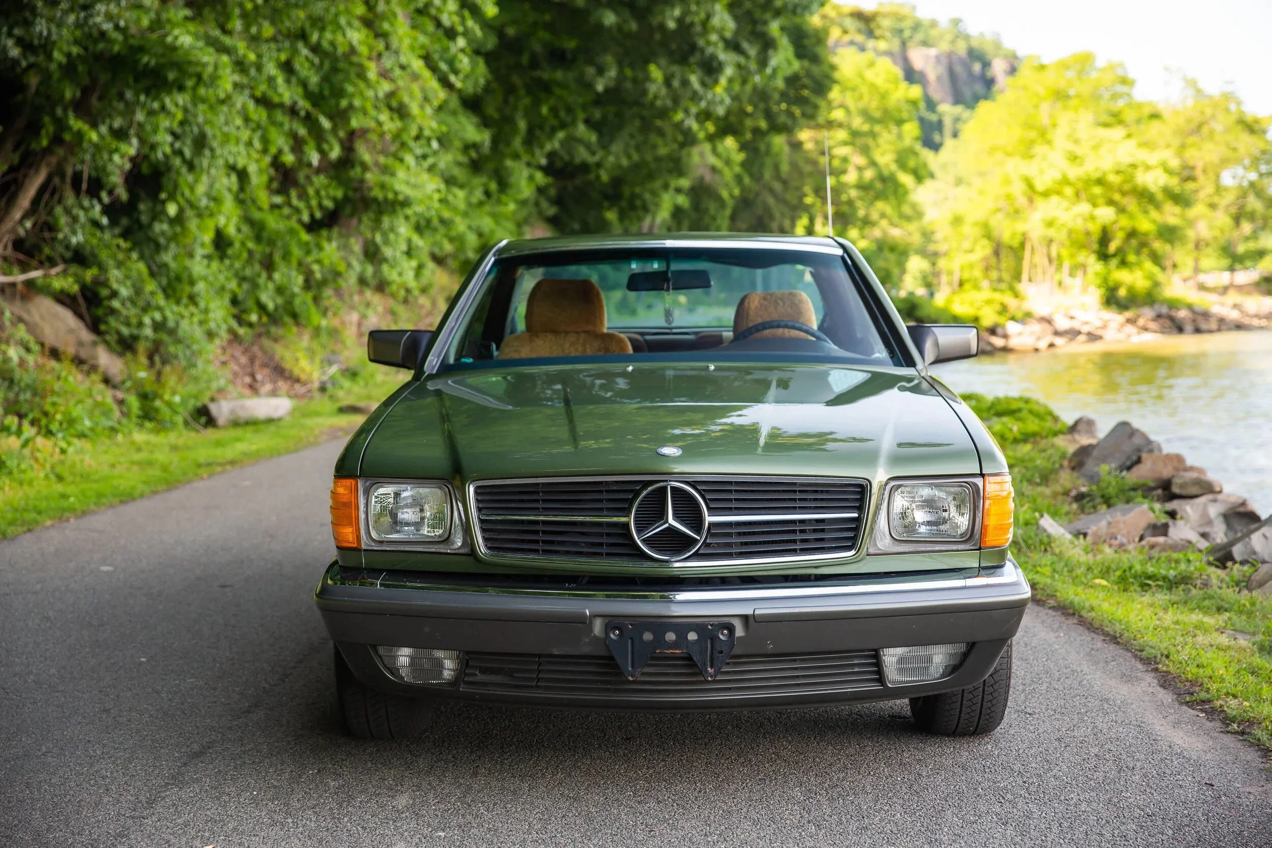 Front view of a green vintage Mercedes-Benz car parked on a scenic road next to a body of water, surrounded by green trees.