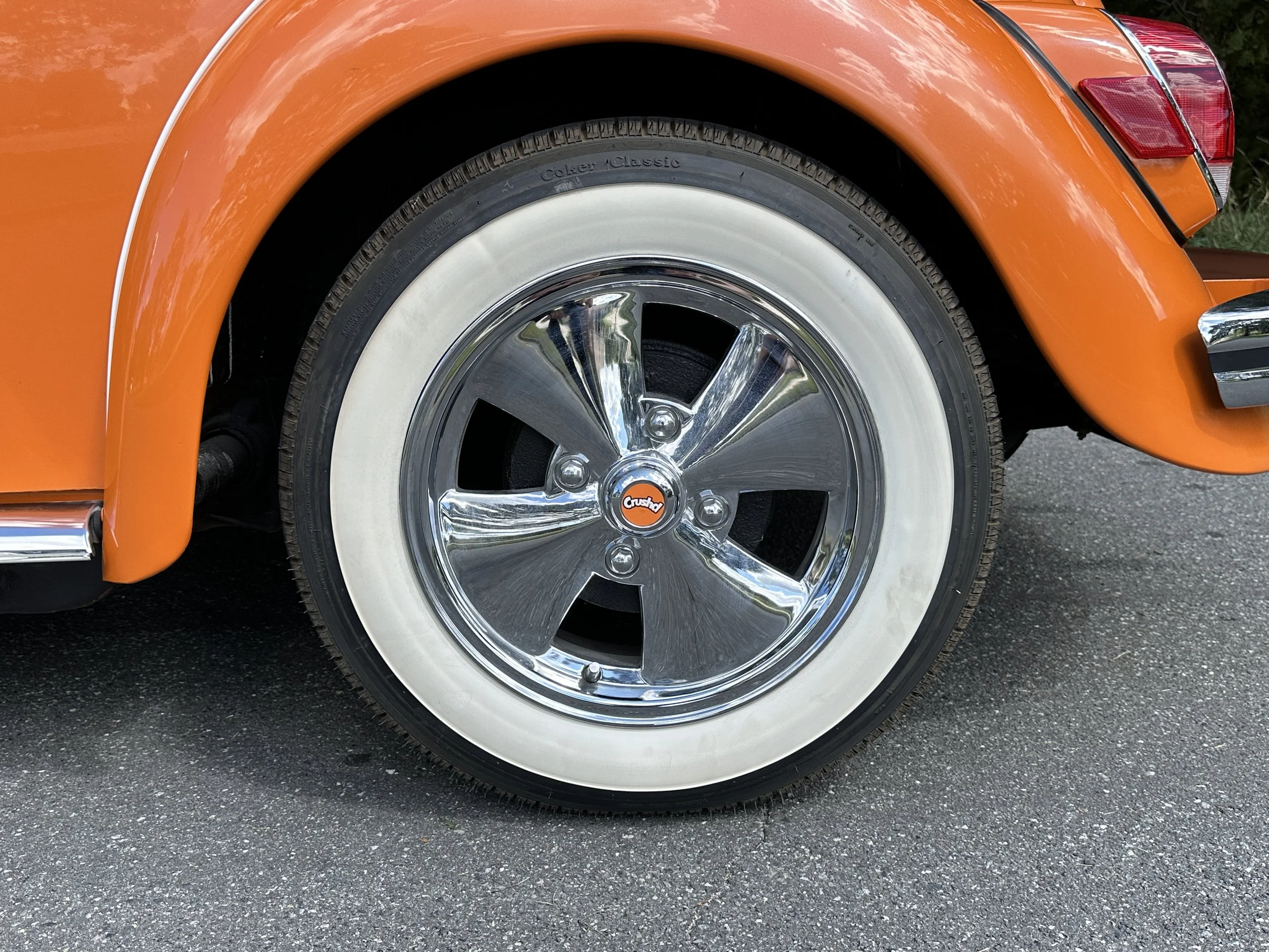 Close-up image of a classic orange car's front wheel with shiny chrome hubcap, white sidewall tire, and part of the orange fender and headlight.