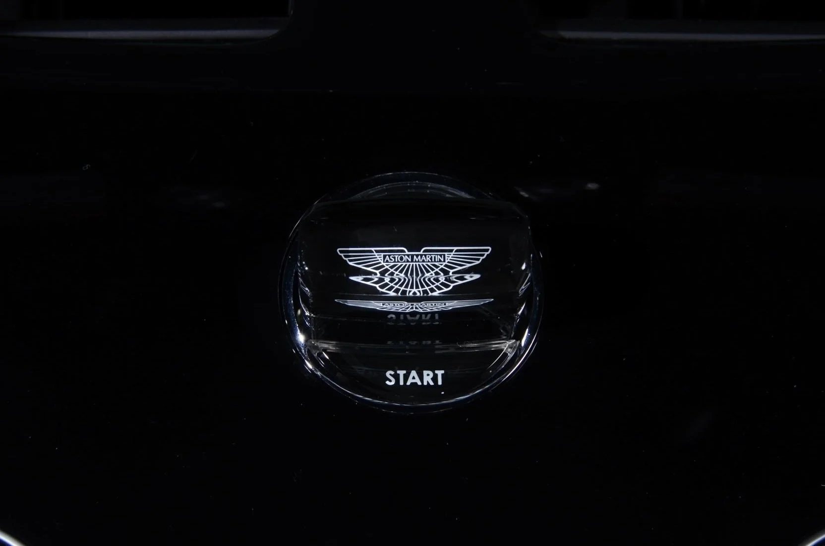 A close-up of a car start button with the Aston Martin logo illuminated in white, surrounded by black surface.