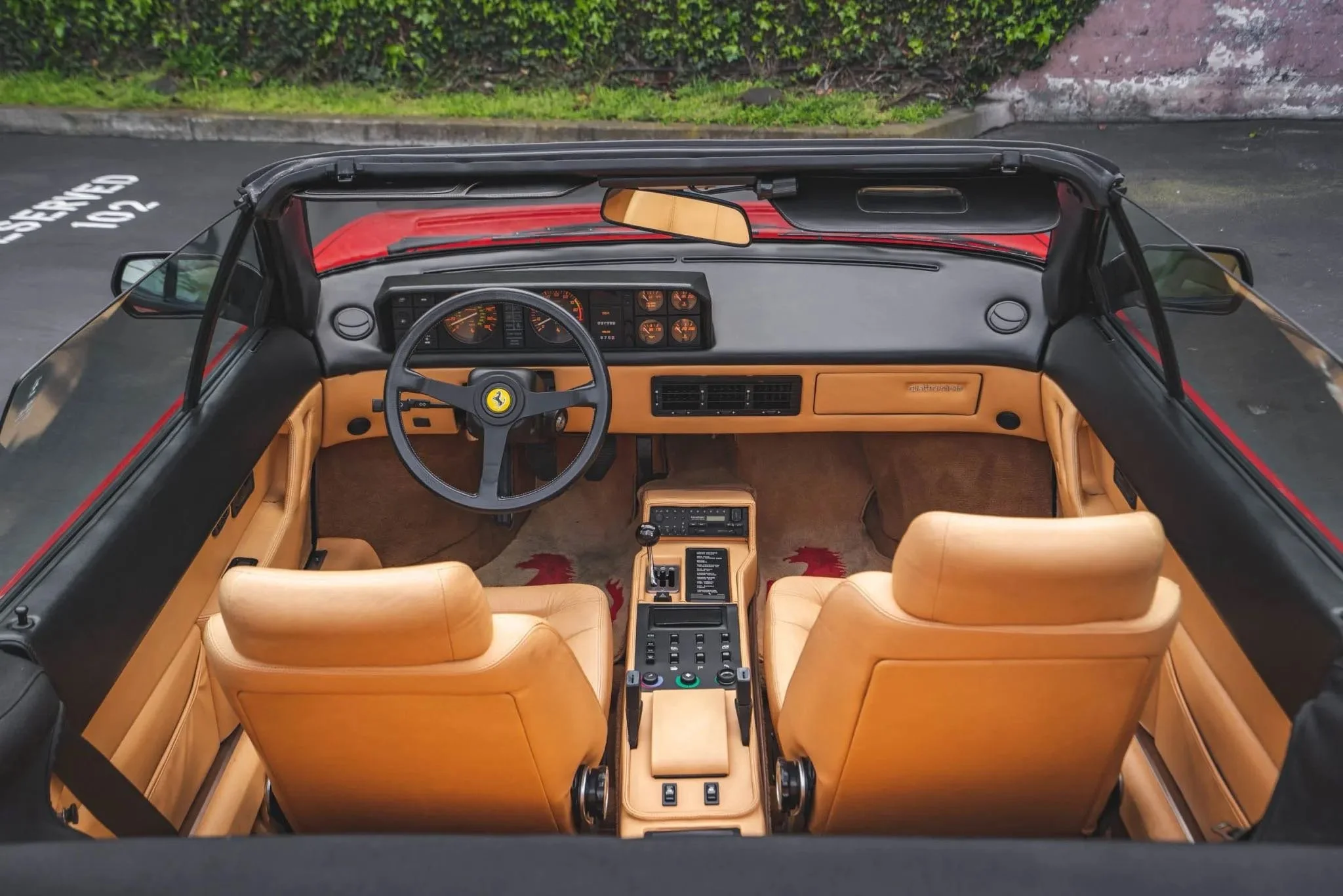 Interior view of a classic Ferrari convertible sports car showing tan leather seats, dashboard with gauges, and a three-spoke steering wheel with Ferrari logo.