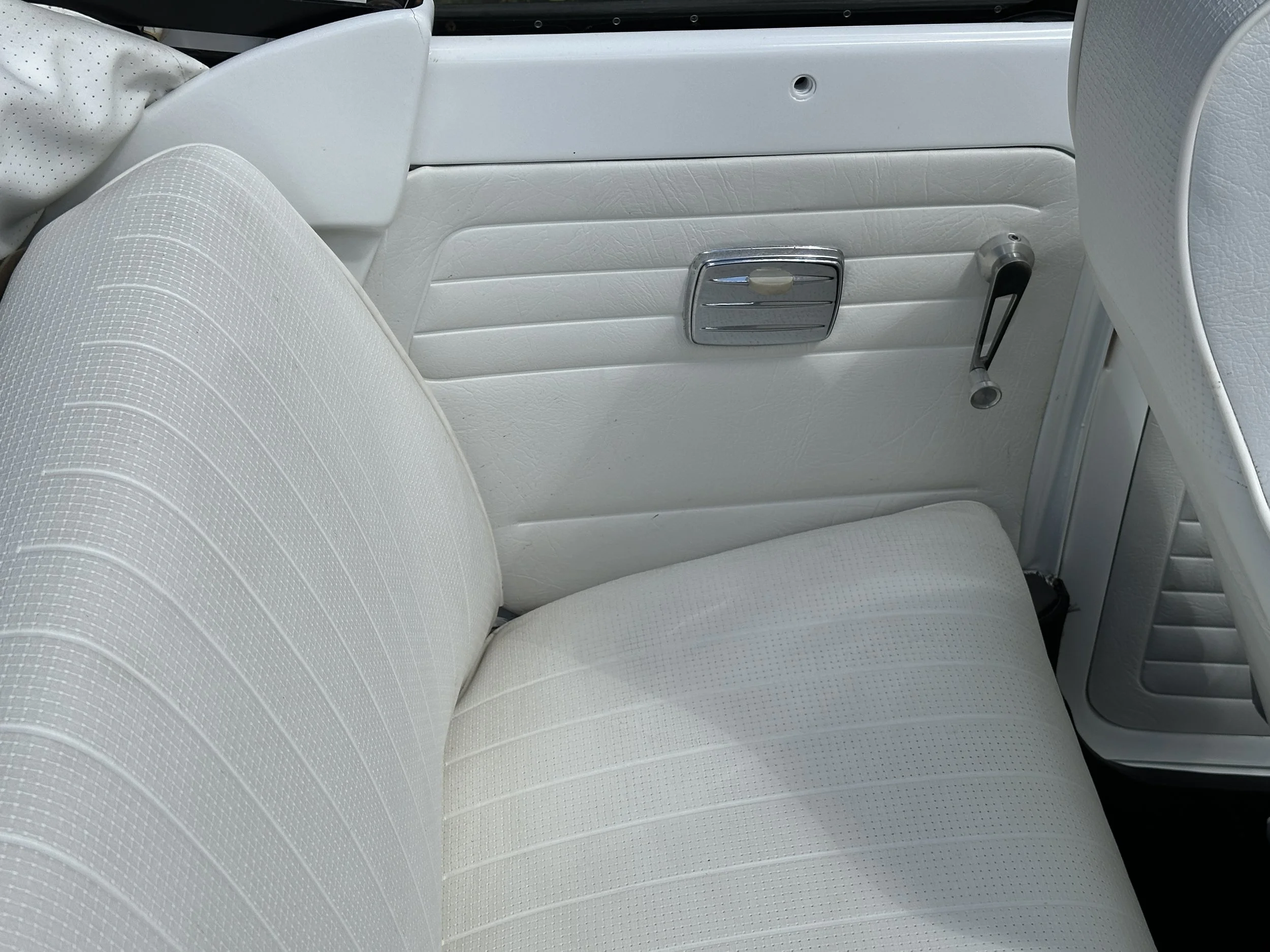 A close-up of a white vintage car interior, showing a white upholstered bench seat, a door panel with a chrome handle, and a window crank.