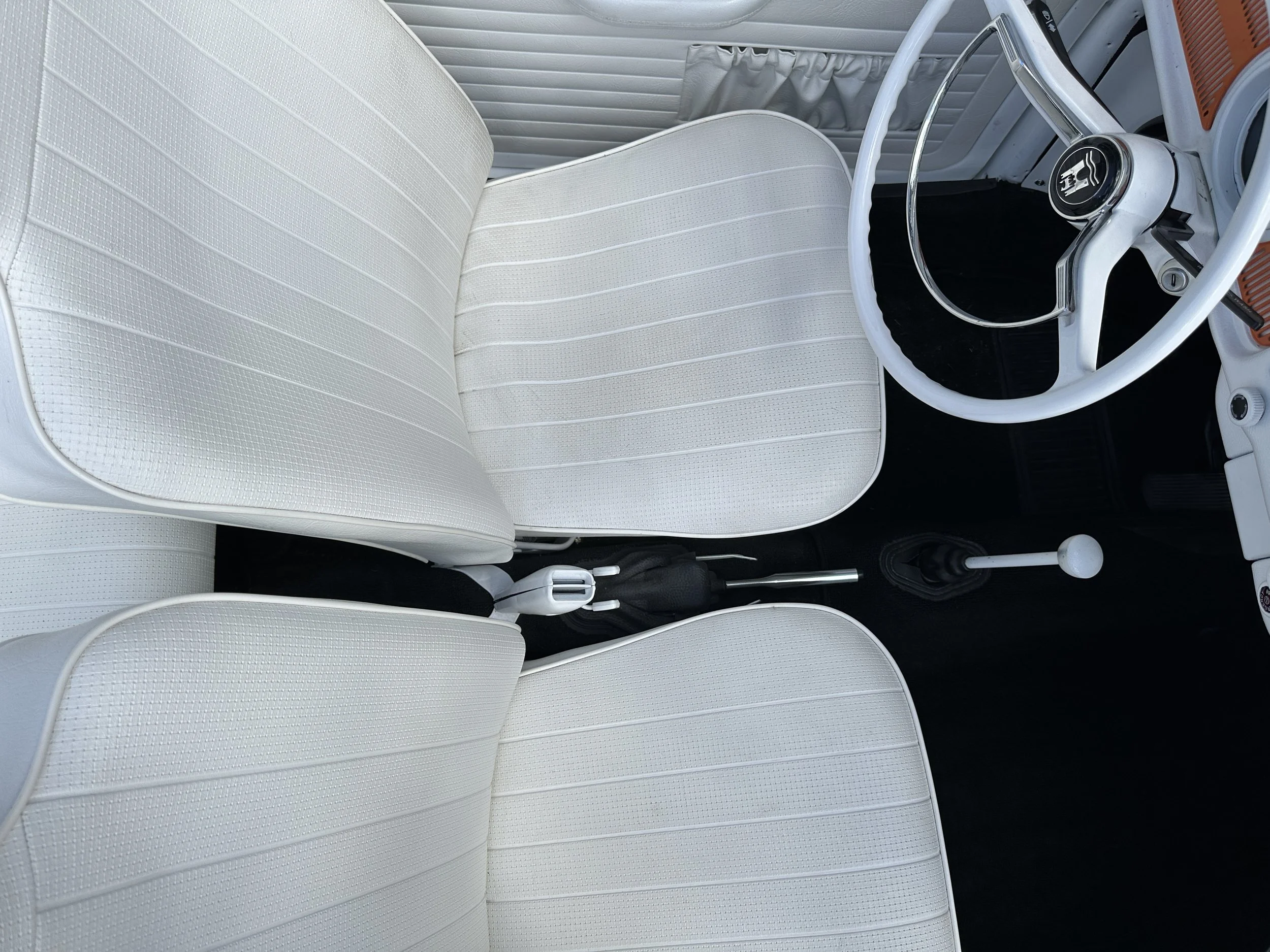 Interior of a vintage car with two white upholstered front seats, a white steering wheel, and a gear shifter, with a view of the passenger side door and dashboard.