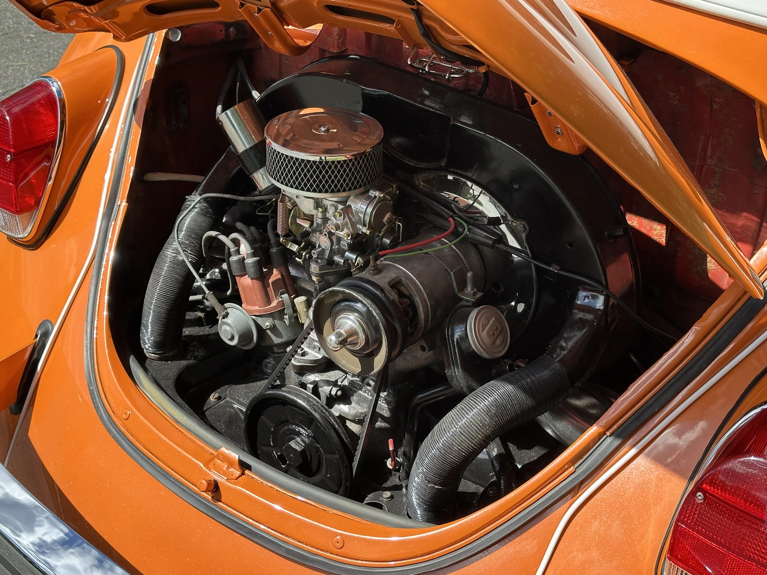 Close-up view of the engine compartment of an orange vintage car, showing parts such as a carburetor with a round air filter, belts, hoses, and other engine components.