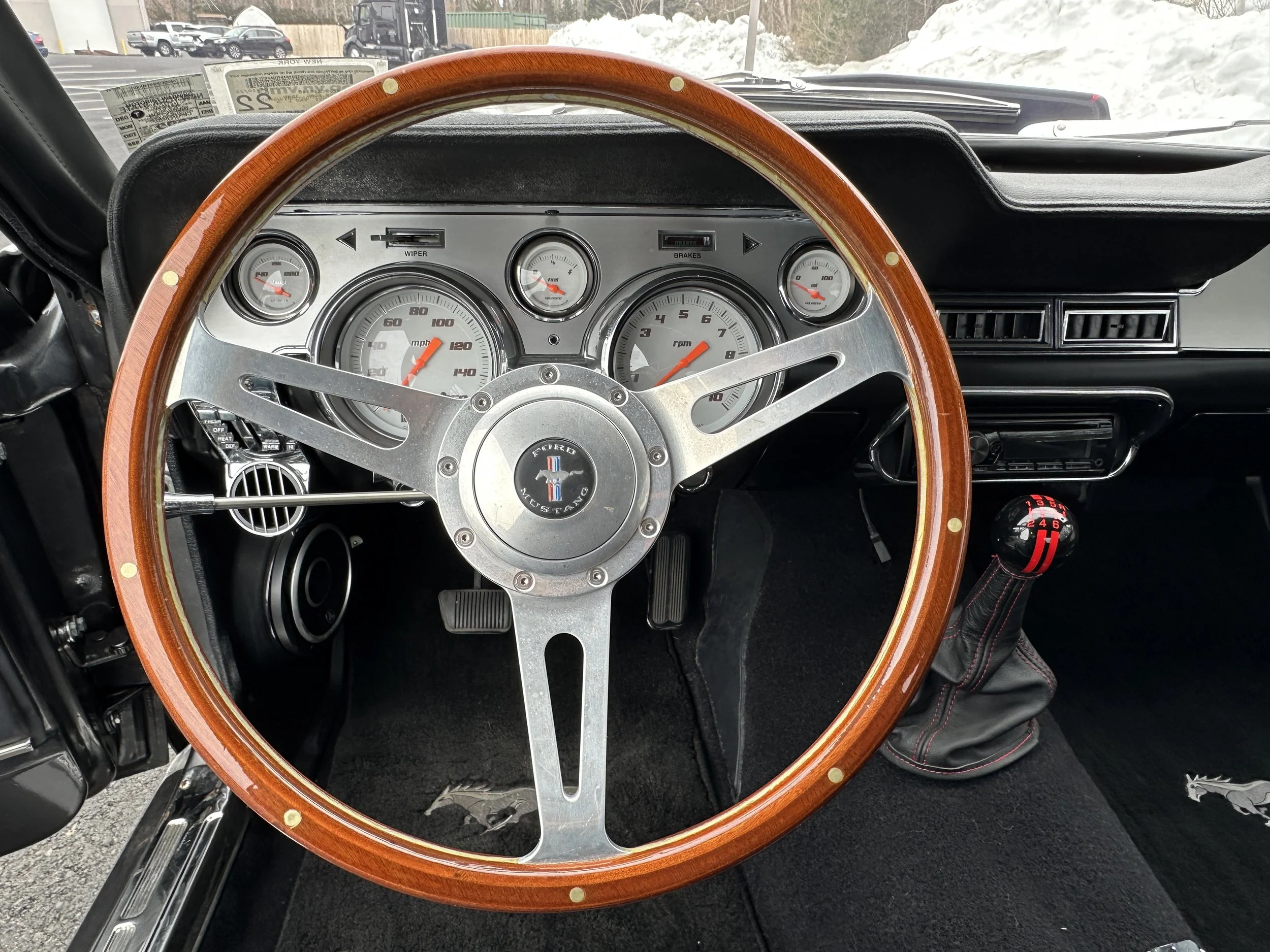The interior of a classic Ford Mustang, featuring a wood-rimmed steering wheel, analog gauges including speedometer and tachometer, a manual gear shift with red accents, and a black dashboard with air vents.