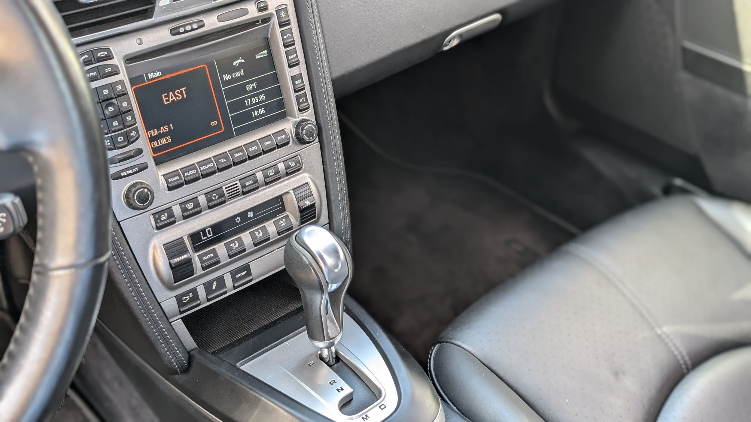 The interior of a car showing the center console with an automatic gear shift, climate control buttons, and a digital display on the dashboard indicating a radio station, temperature, date, and time.