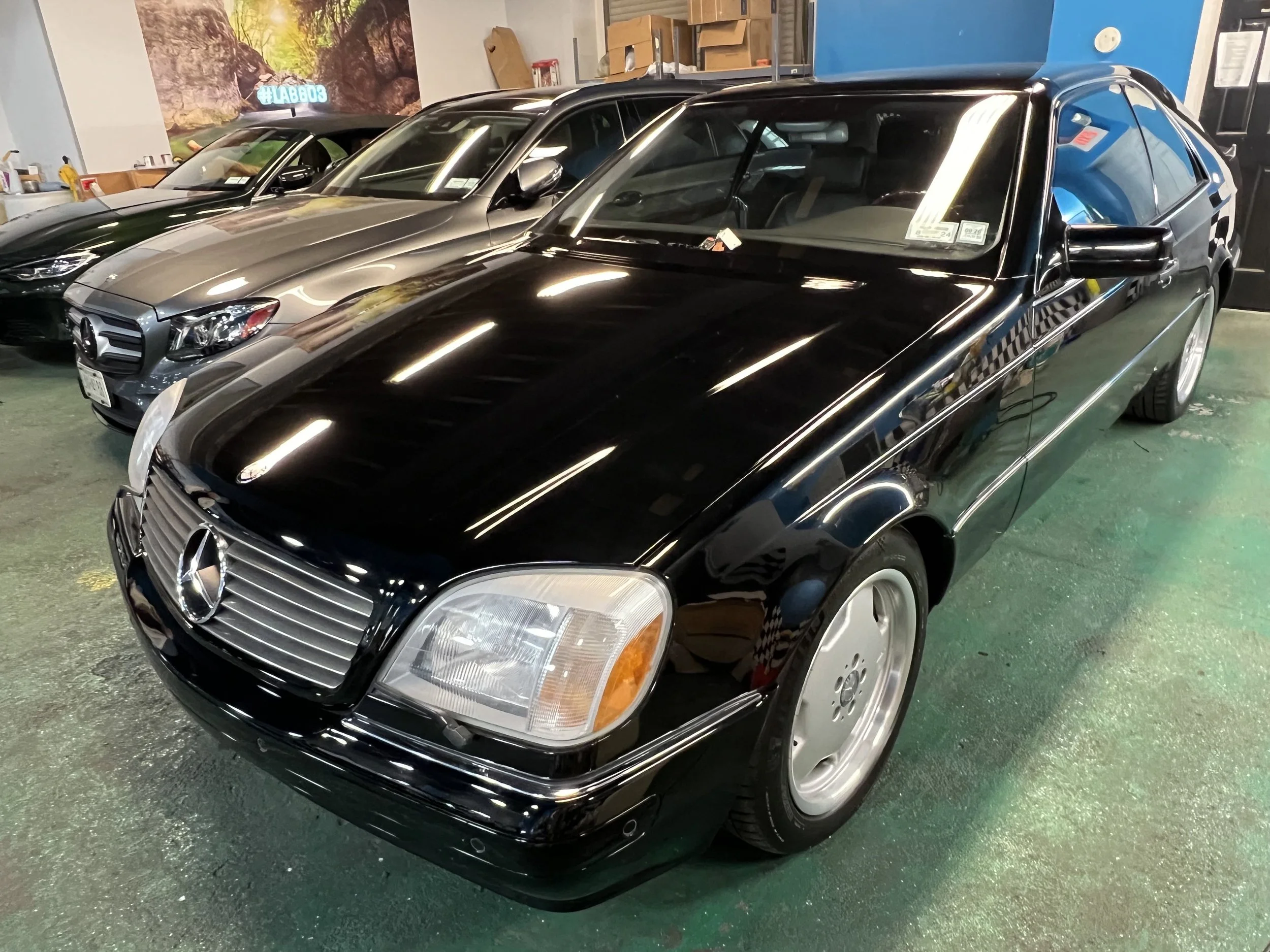 Black vintage Mercedes-Benz coupe parked indoors among other cars, with a shiny, reflective surface.