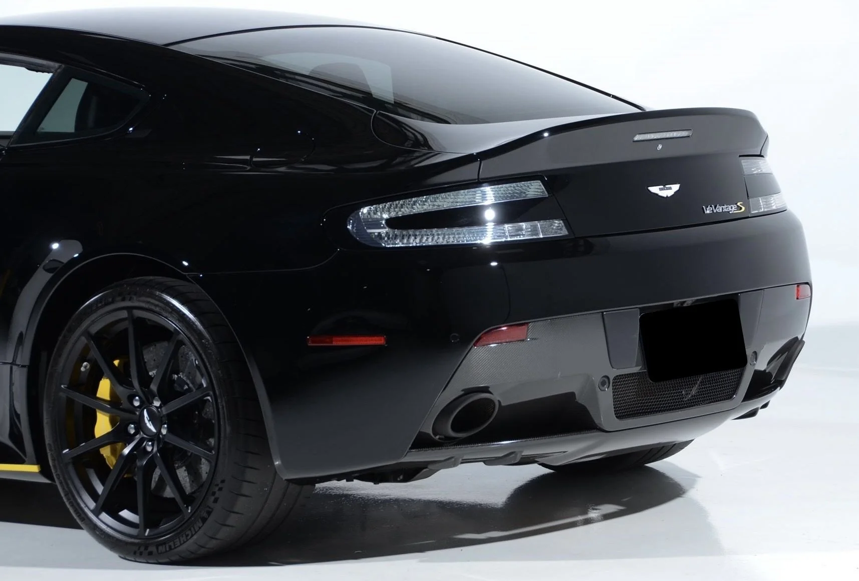 Front rear view of a black Aston Martin V12 Vantage S sports car with a sleek aerodynamic design, black alloy wheels with yellow brake calipers, and carbon fiber accents.