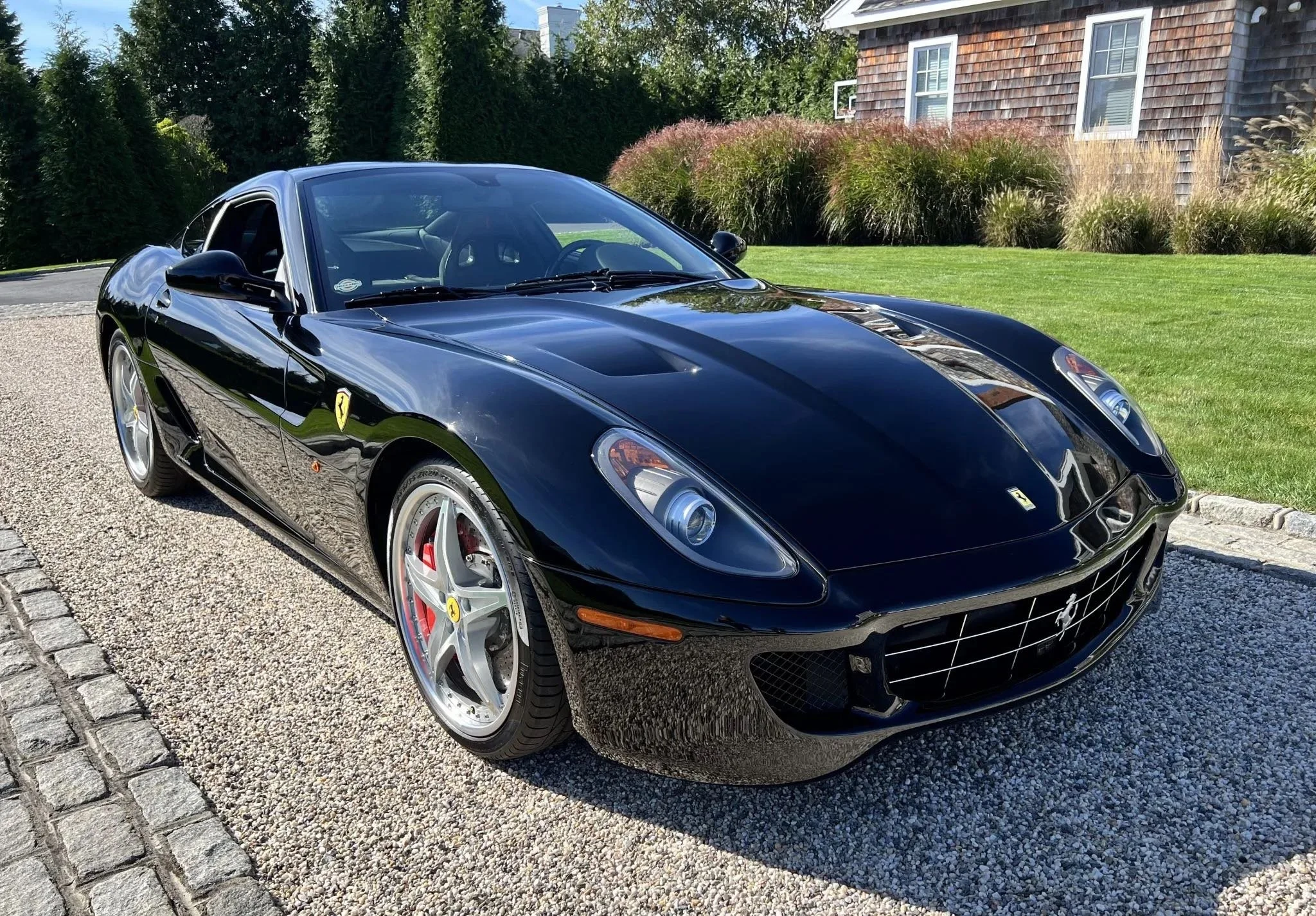 Black Ferrari sports car parked on a gravel driveway with a landscaped background including a lawn, bushes, and a wooden house.