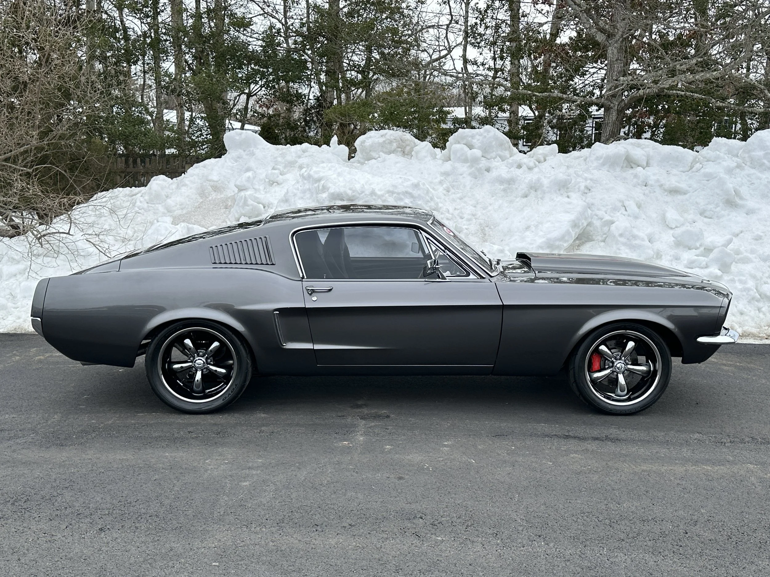A classic gray muscle car parked on asphalt with snow mounds and trees in the background.