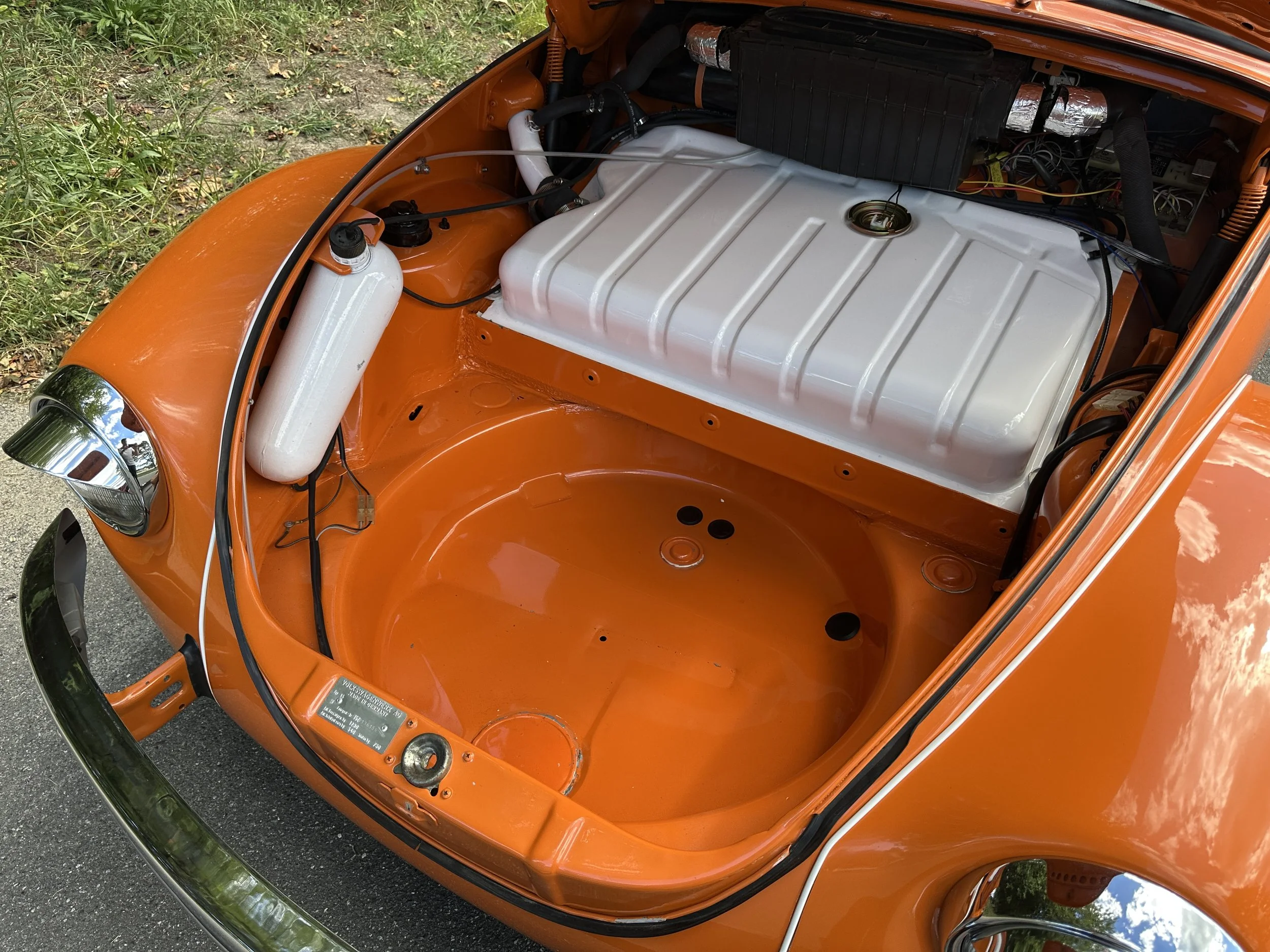 Orange Volkswagen Beetle with engine bay open, showing empty space where the engine goes, a white fuel tank, and various mechanical components.