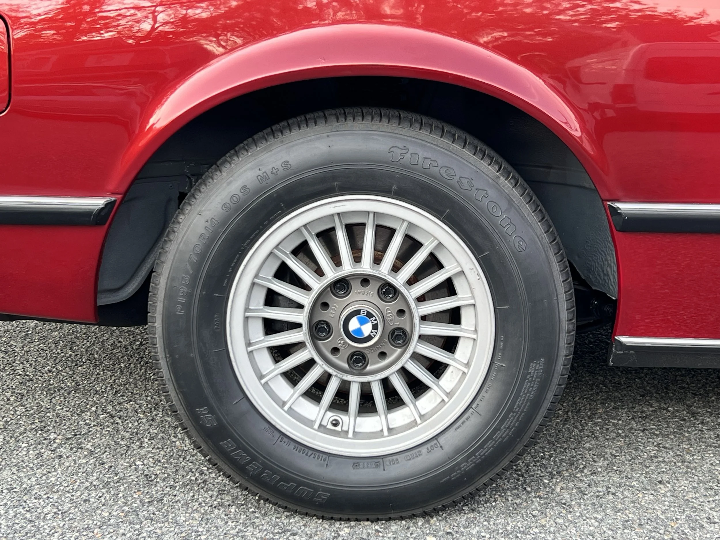 Close-up of a classic red BMW 630 csi front wheel with a BMW emblem on the hubcap, tire marked 'Firestone' and 'S-9594,' and the car's red body and black trim visible.