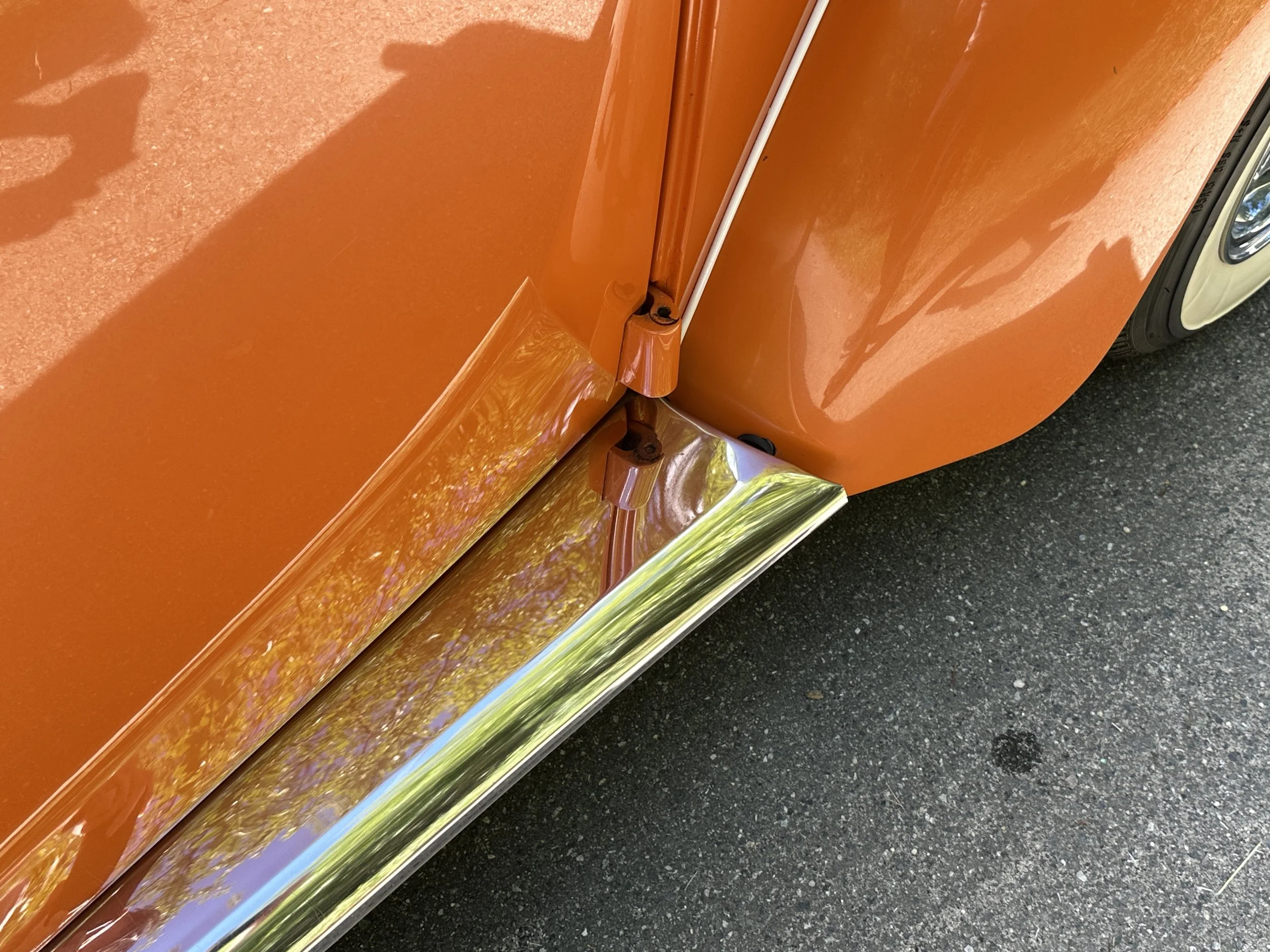 Close-up of the front fender and door of an orange vintage car, showing shiny chrome trim and part of the black tire.