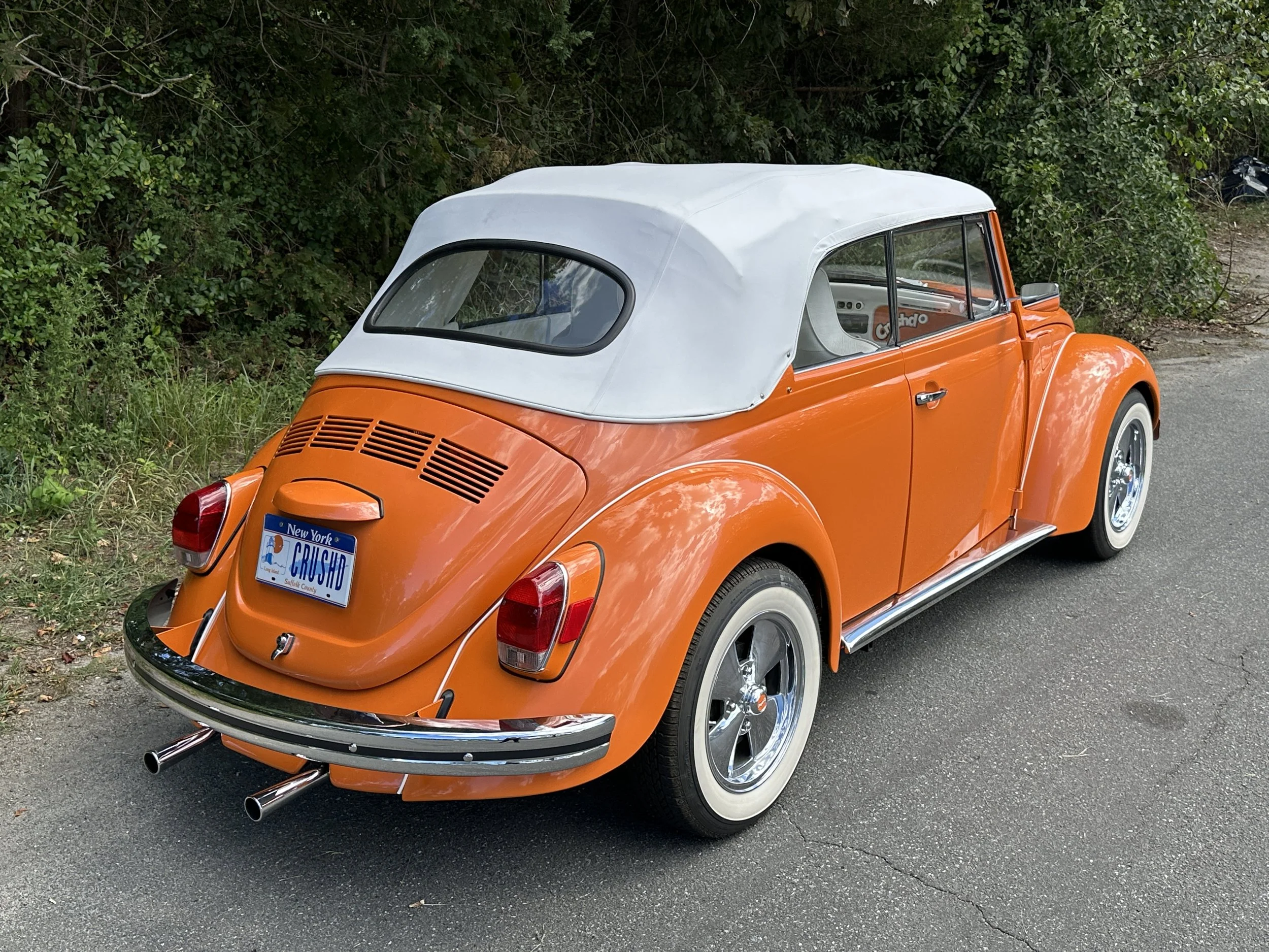 Orange vintage Volkswagen Beetle convertible with white soft top parked on the side of a road, greenery in background.