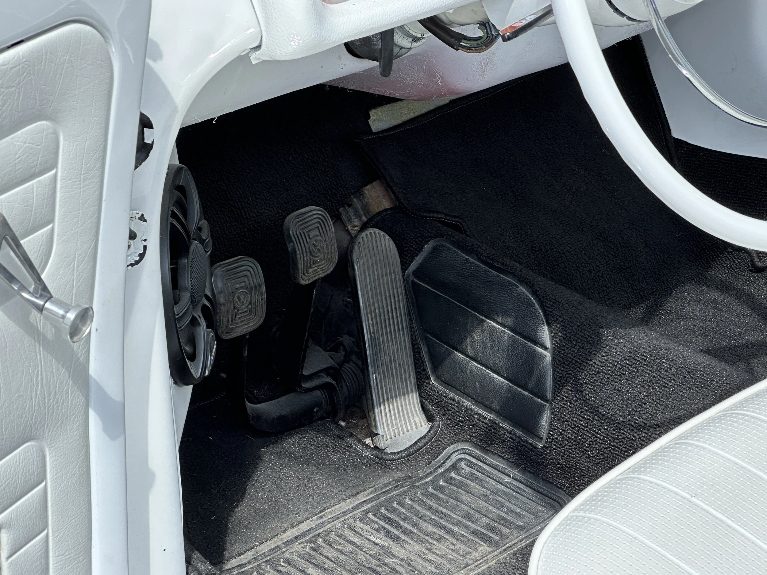 Interior of a vintage car showing three pedals, a white steering wheel, black floor mats, and a black dashboard with speaker system.