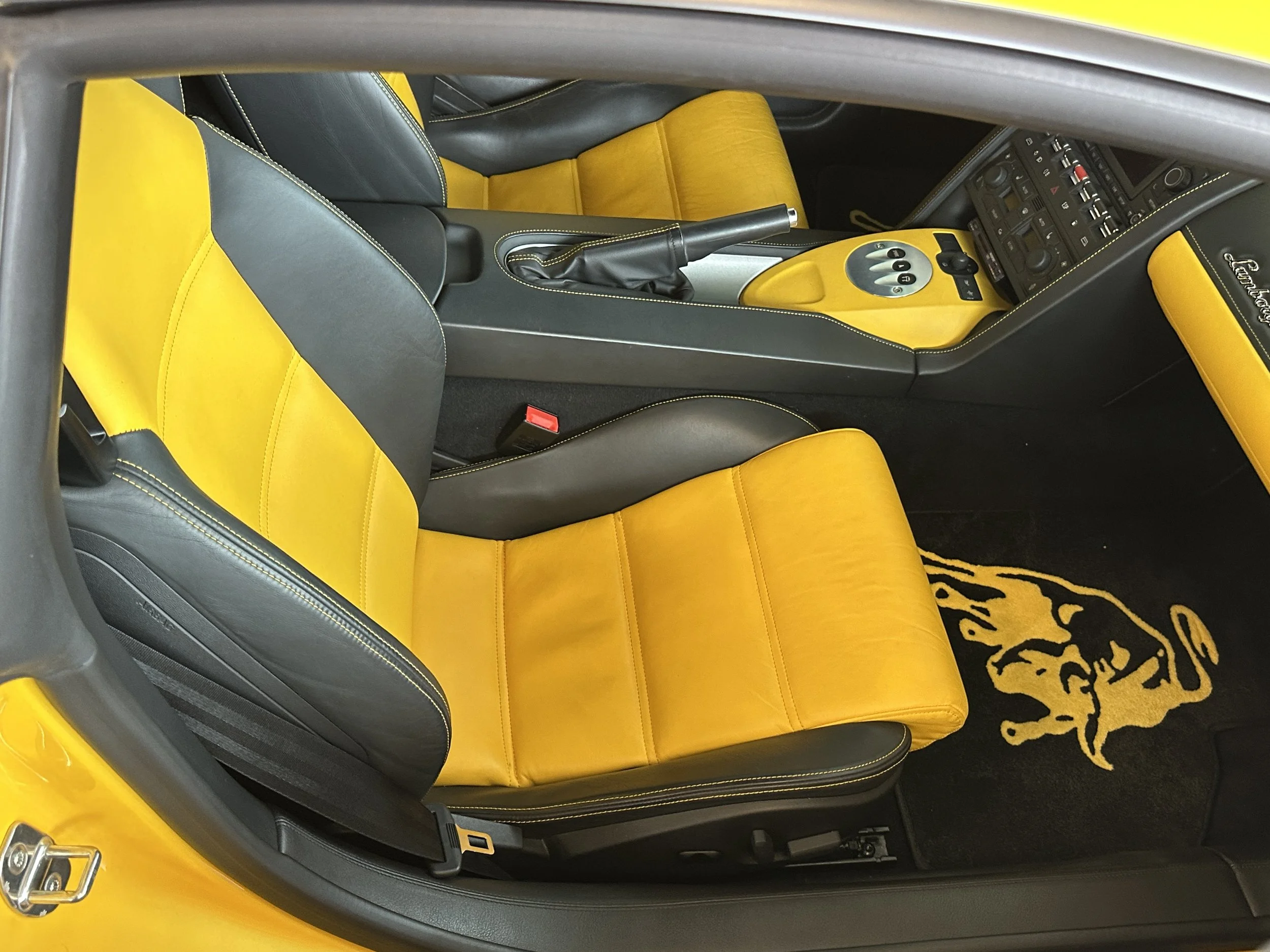 Interior of a yellow sports car, showing yellow and black leather seats, black center console, and dashboard with various controls. There is a black floor mat with a yellow lion logo.