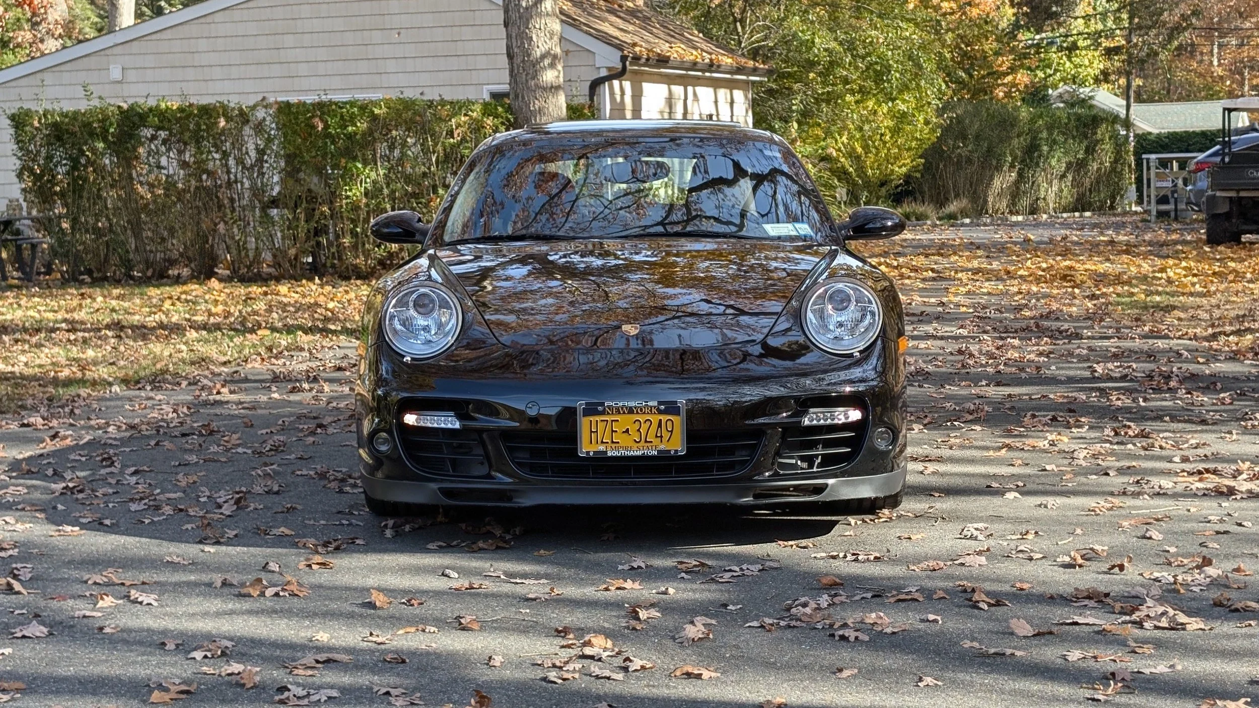 Black Porsche sports car parked on a driveway, with fallen autumn leaves and trees in the background.