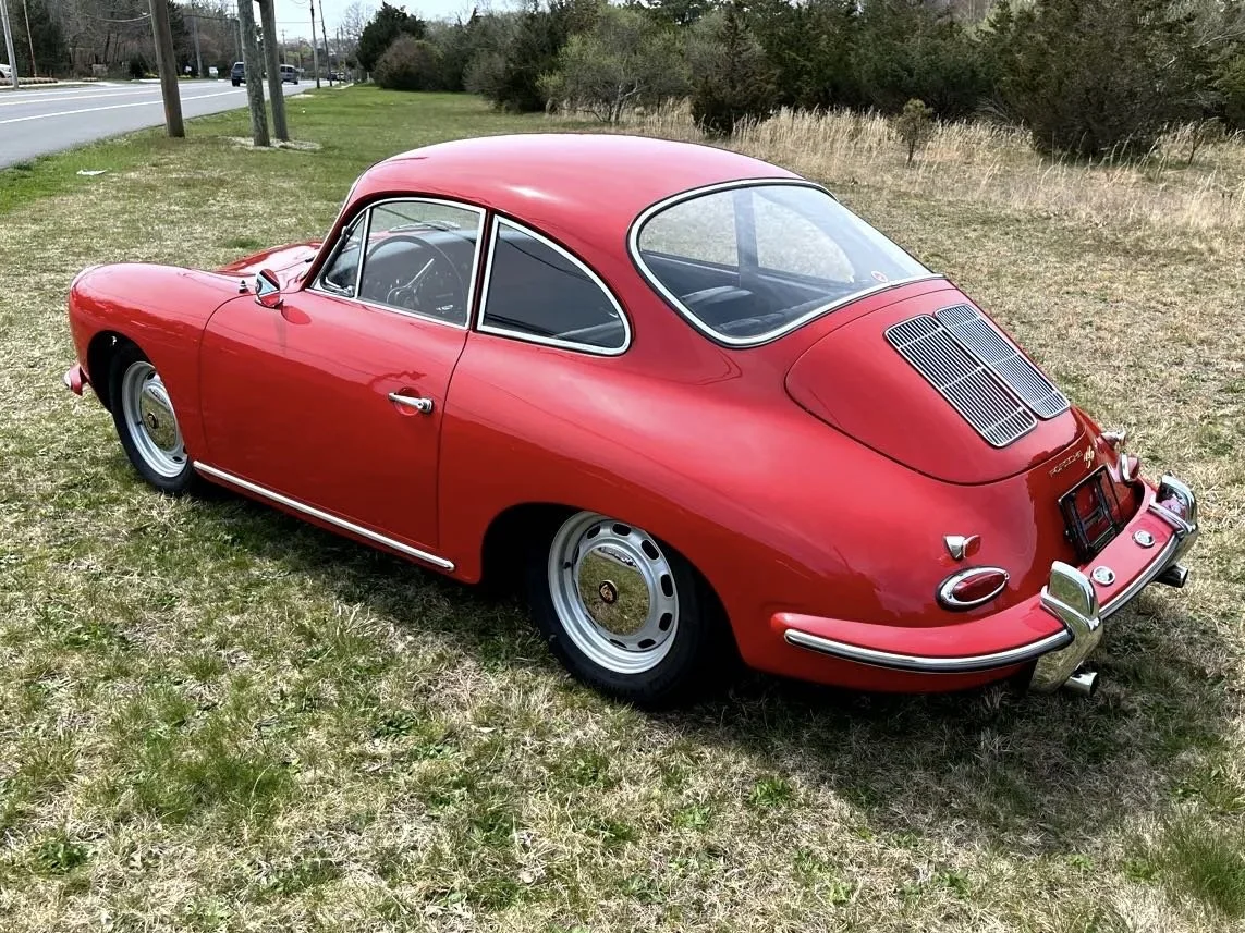 A red vintage Porsche 356 sports car parked on a grassy area near a road, viewed from the rear and side.