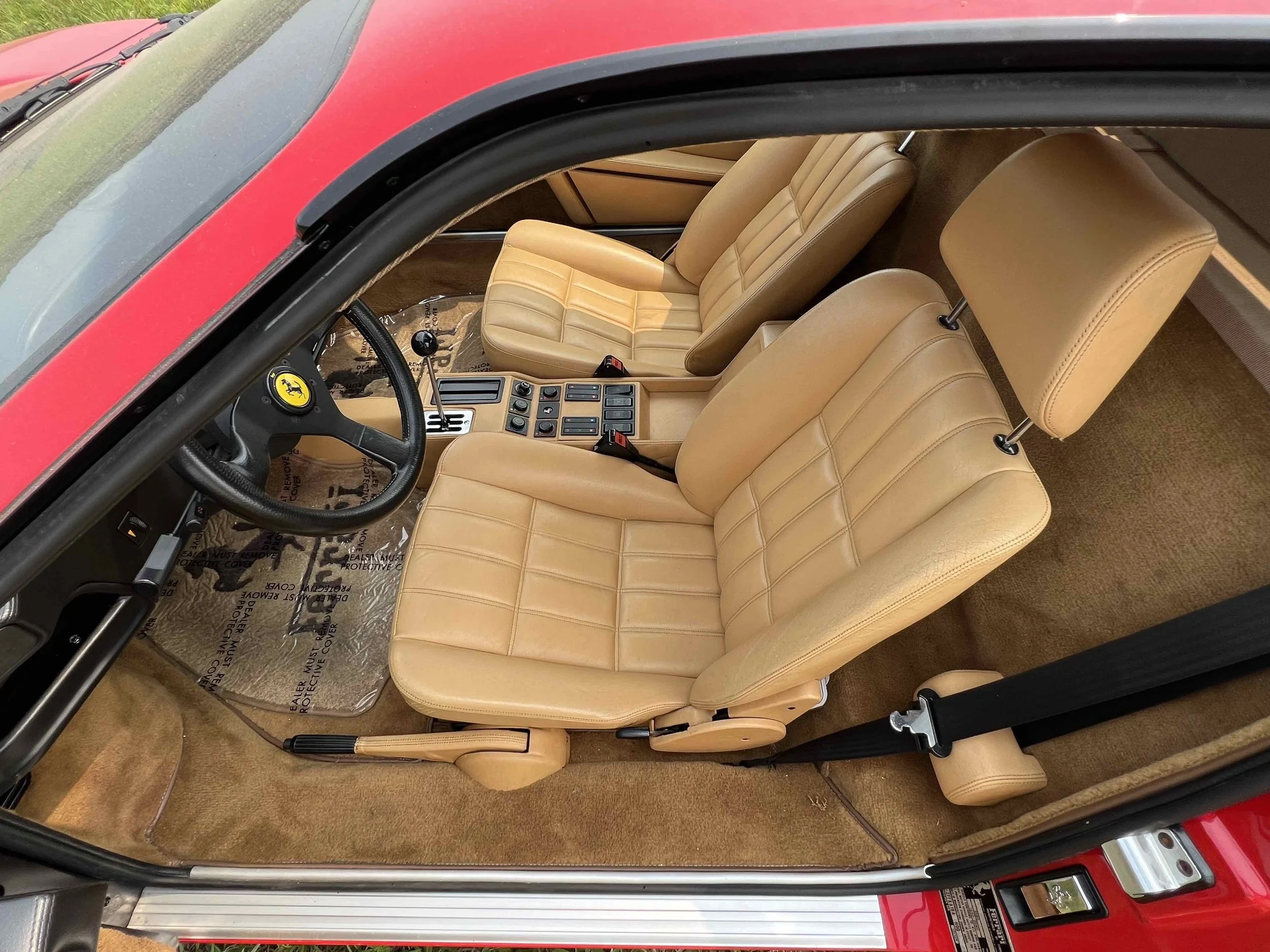 Inside of a classic Ferrari car with tan leather seats, a black steering wheel with Ferrari logo, and beige carpeting.