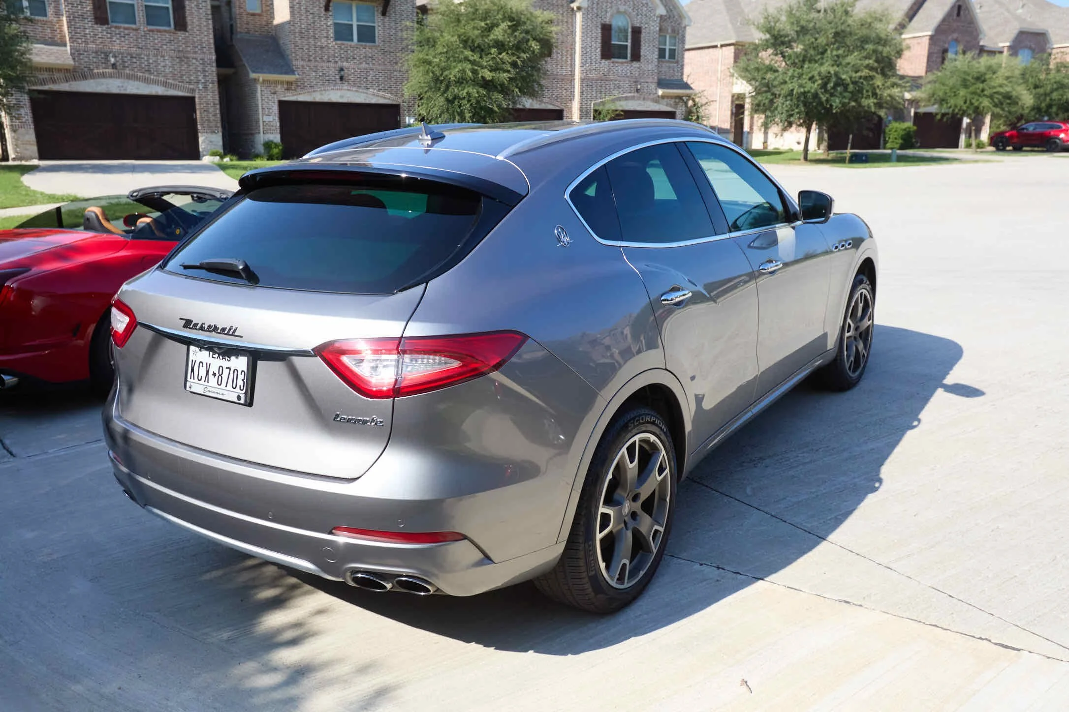 A silver Maserati Levante SUV parked in a driveway with a red sports car nearby and residential houses in the background.
