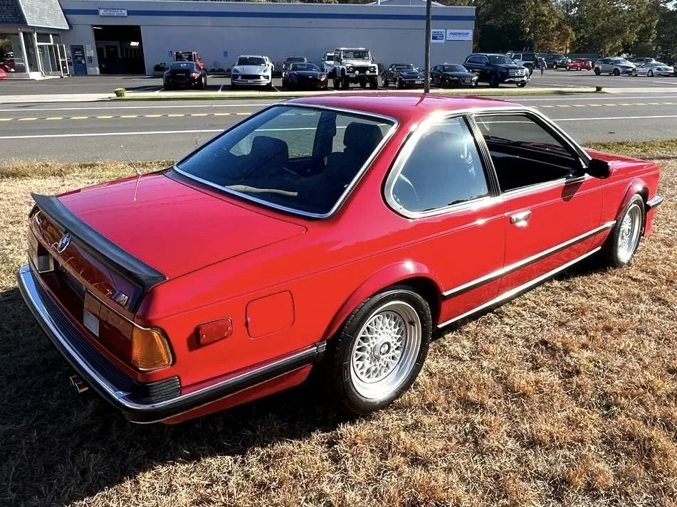 A red vintage BMW coupe parked on a grassy area near a busy street with cars and a commercial building in the background.
