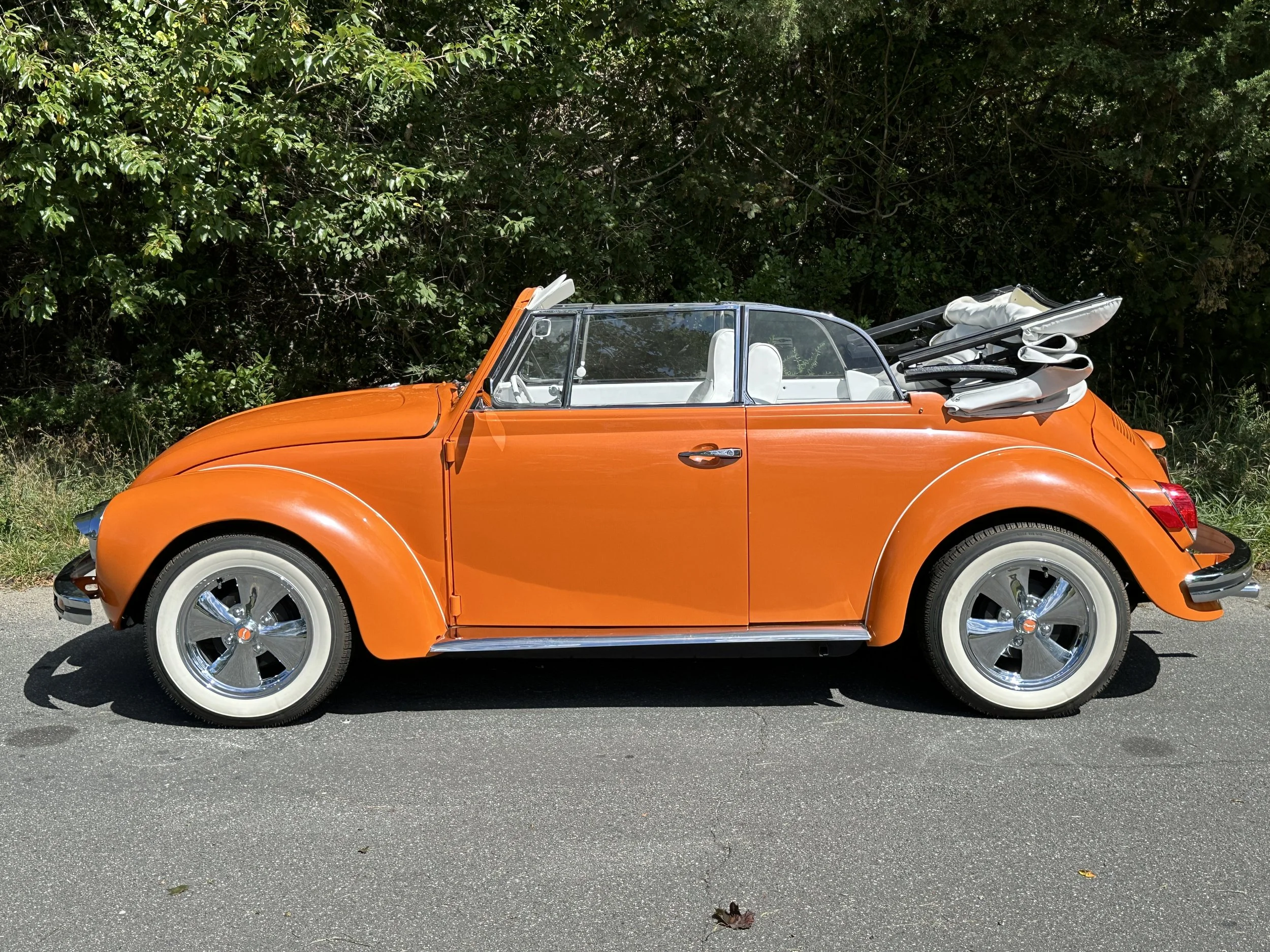 Orange vintage Volkswagen Beetle with convertible top folded back, parked on the side of a road with green foliage in the background.