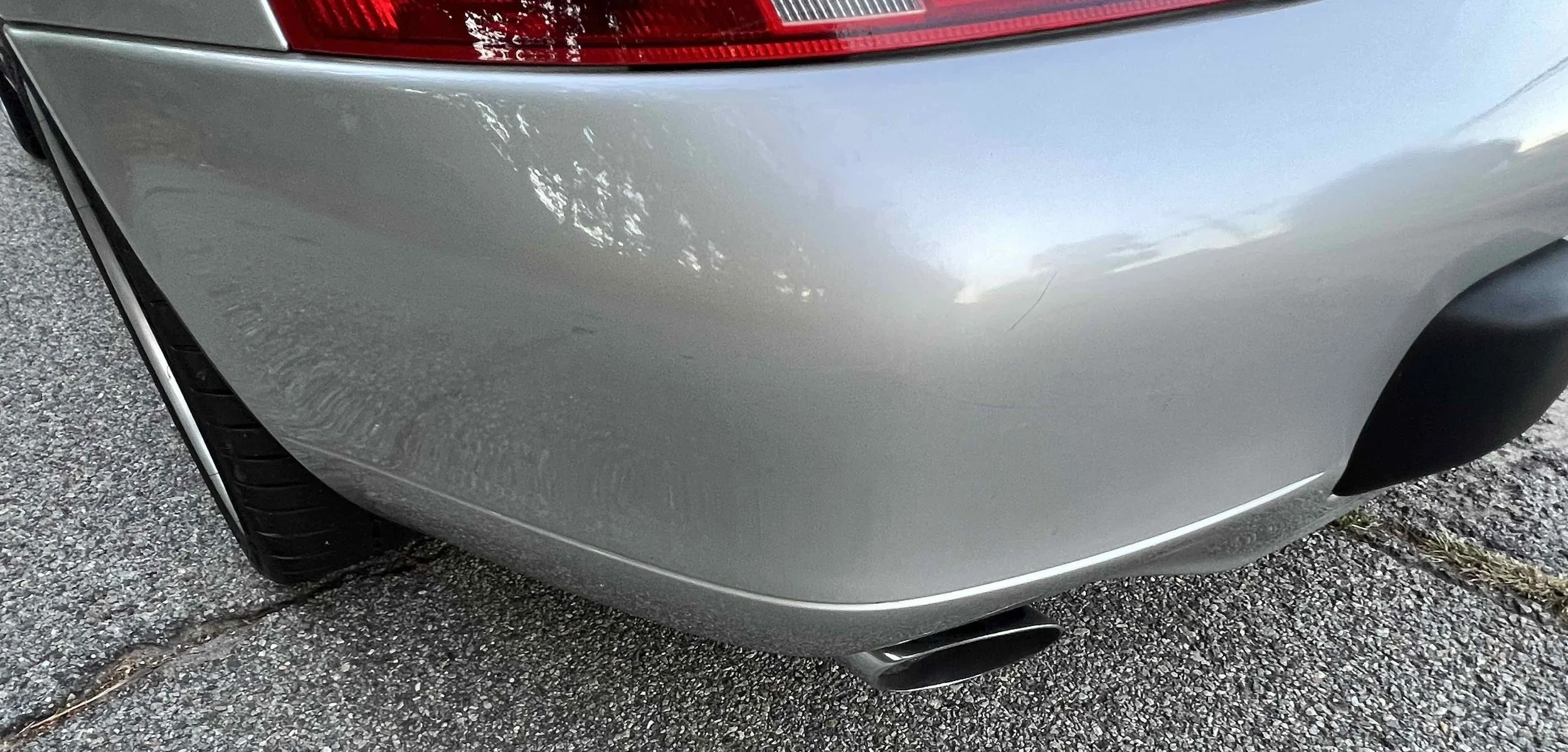 Close-up of the rear end of a silver car, showing the taillight, part of the rear bumper, a section of the rear wheel, and the exhaust pipe. The car is parked on a cracked asphalt surface.