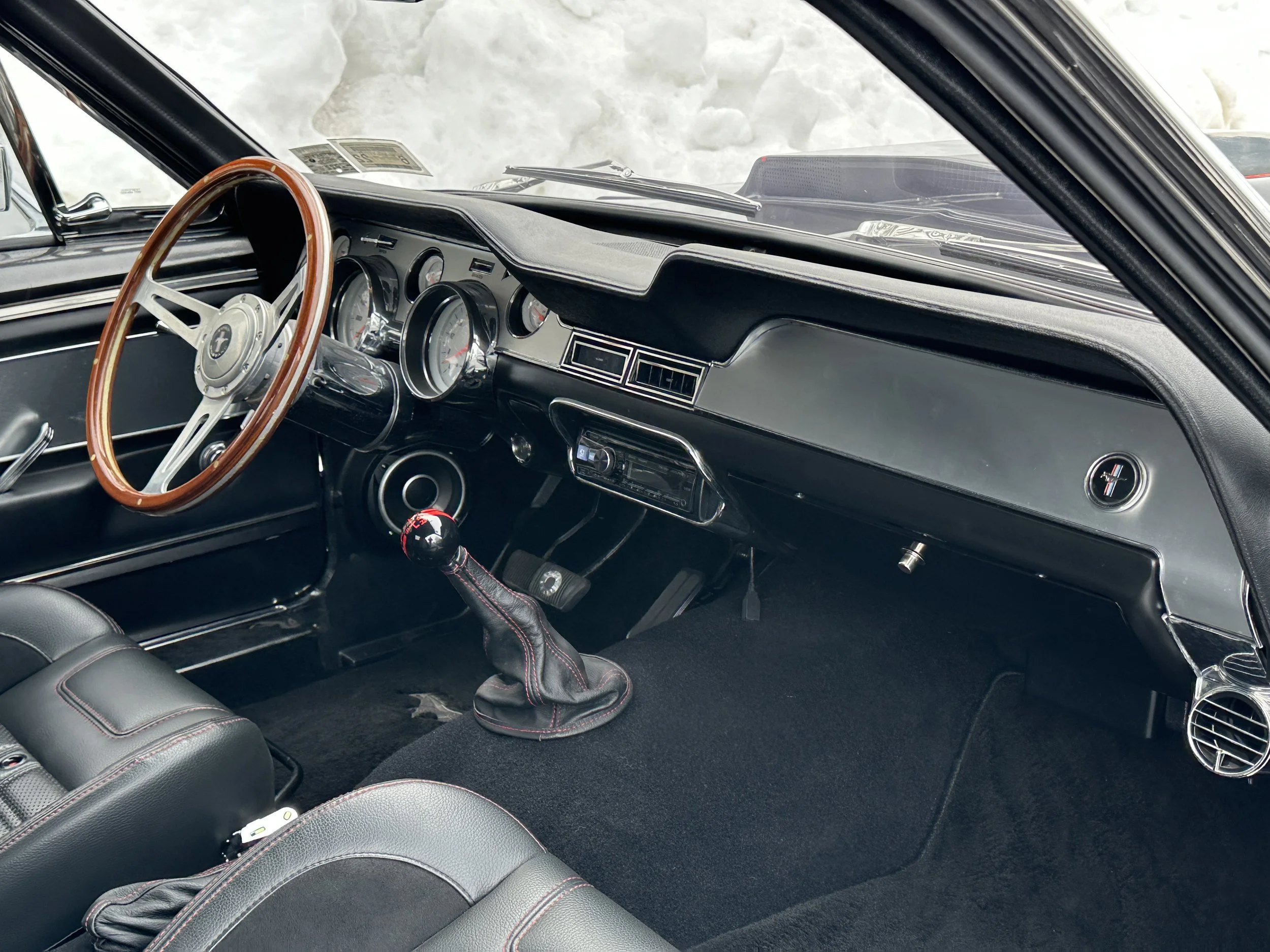 Interior of a classic car showing a wooden steering wheel, analog gauges, a manual gear shift, and black dashboard with a Mustang emblem.