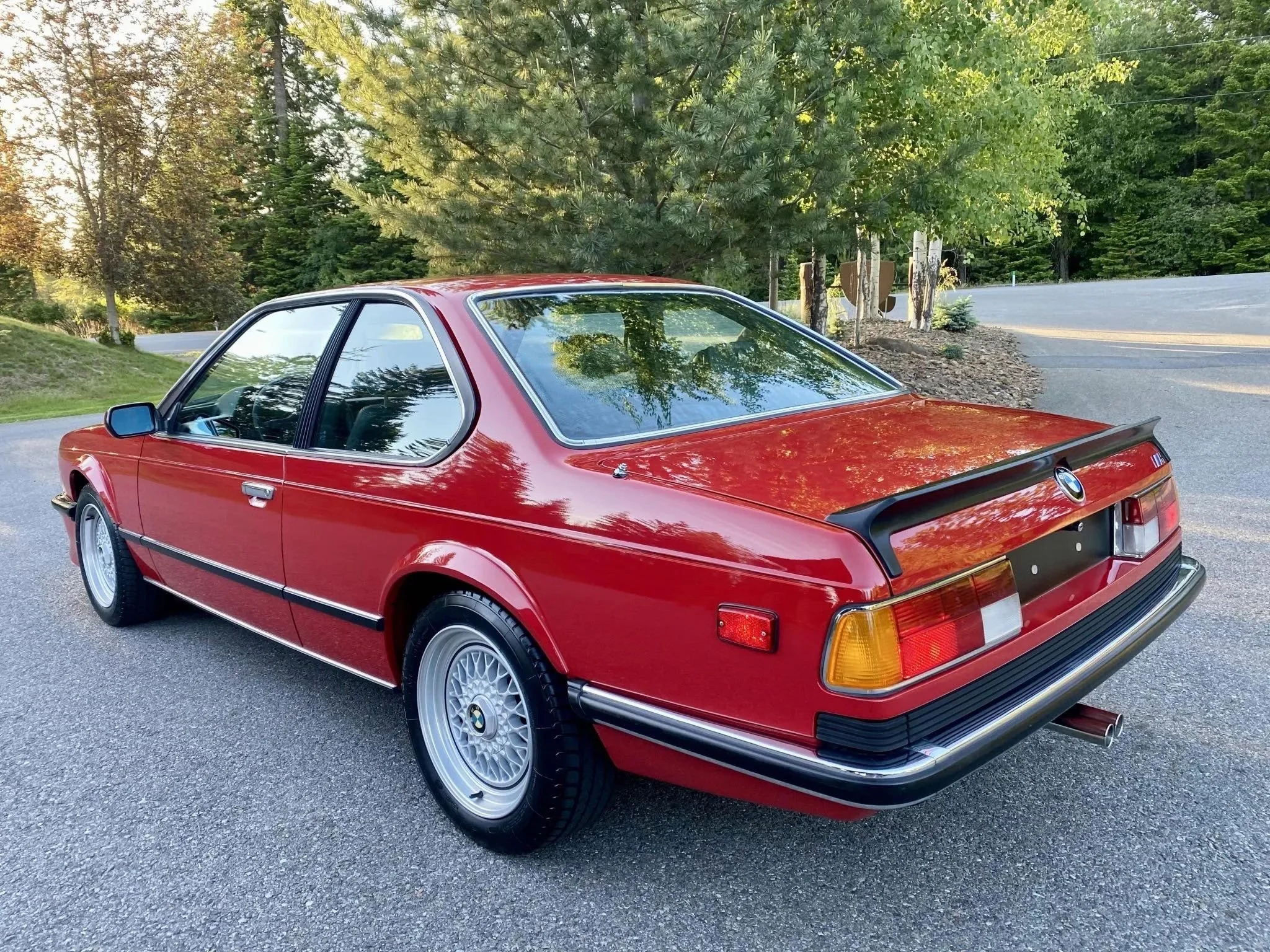 Red vintage BMW coupe car parked on a paved road with trees and greenery in the background.