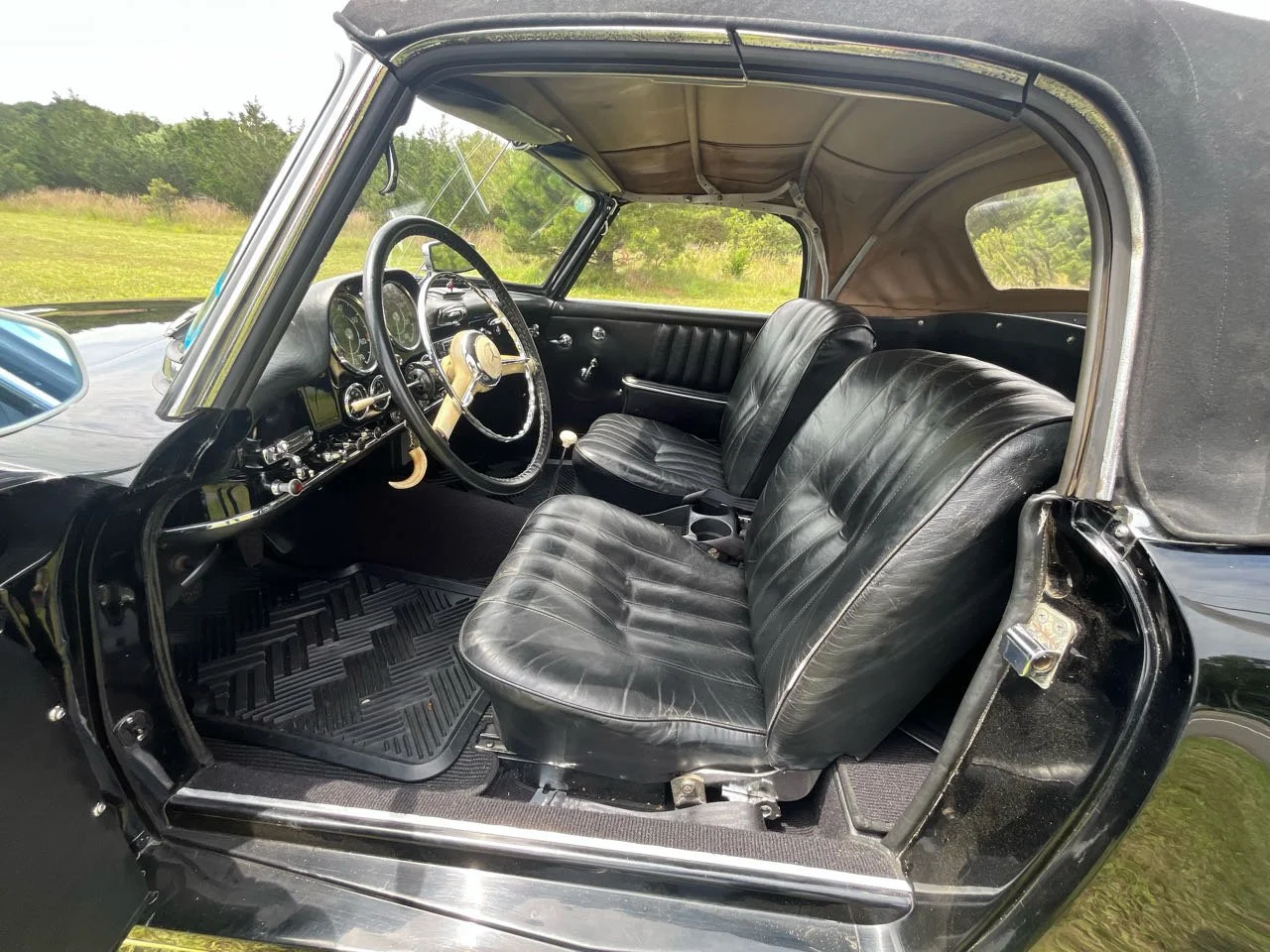 Interior view of a vintage black car showing the dashboard, steering wheel, leather seats, and door panel with window controls. The car has a black fabric roof and is parked outdoors in a grassy area.