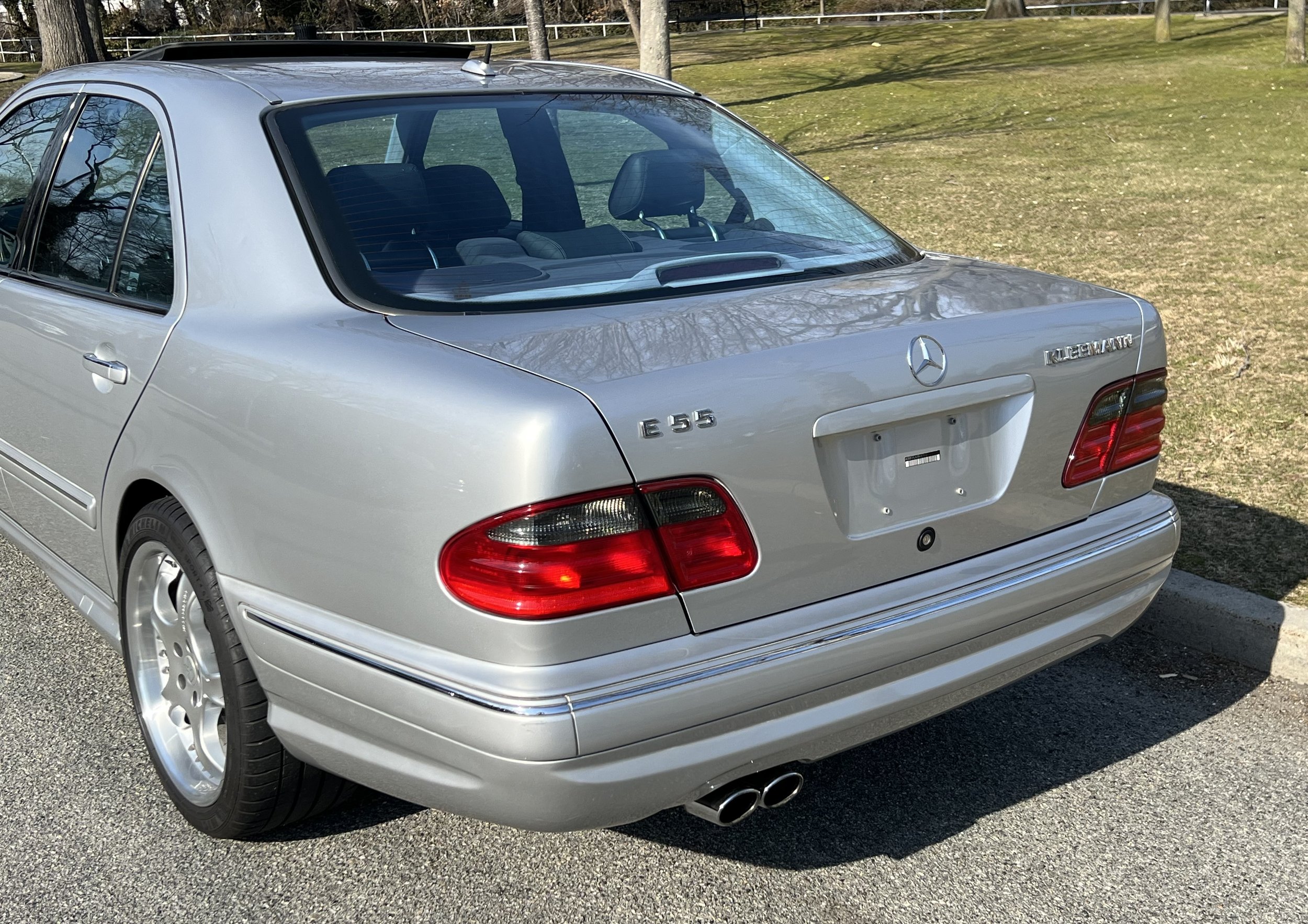Silver Mercedes-Benz E55 AMG sedan parked outdoors on a paved road area with grass and trees in the background, viewed from the rear three-quarters.