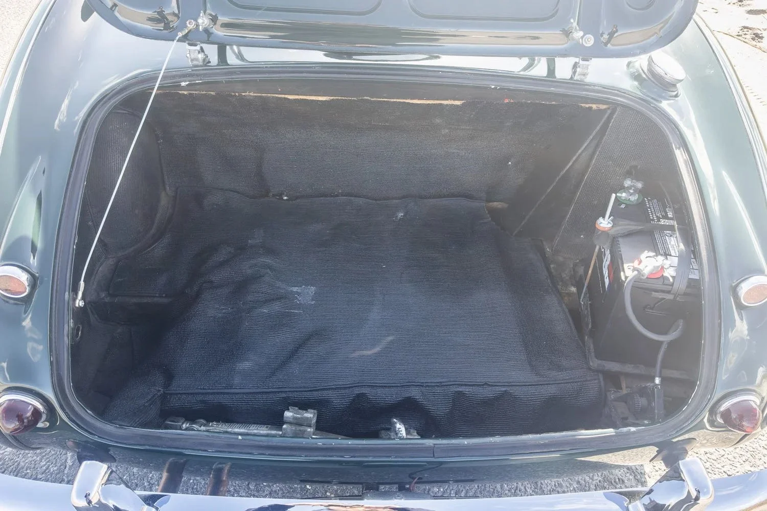 Front trunk of a vintage car with a battery on the right side and a black fabric lining.